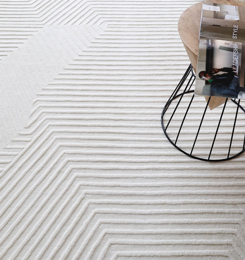 A round wooden table with a wire base sits on the Handmade Decor Area Rug - LKH 012 by RAMSHA HOME, which features ivory geometric, textured patterns; a magazine rests on the tabletop.