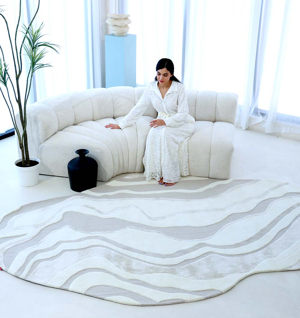 A woman in a long white dress sits on a curved white sofa in a bright room with white curtains, a potted plant, a black vase, and a wavy-patterned rug on the floor.