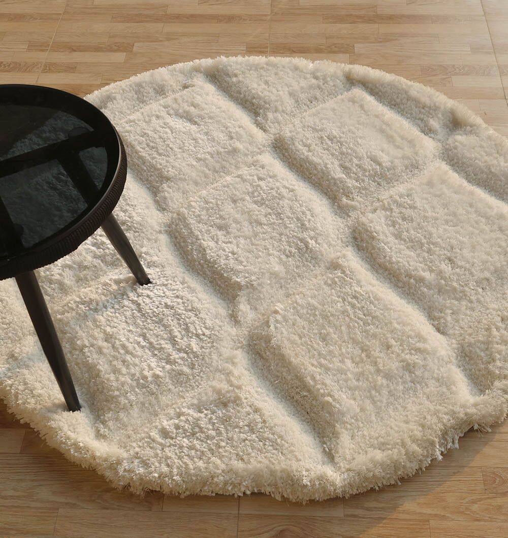 A round, white, textured rug with a raised square pattern is placed on a light wooden floor, partially underneath a black, round-top table with three legs.