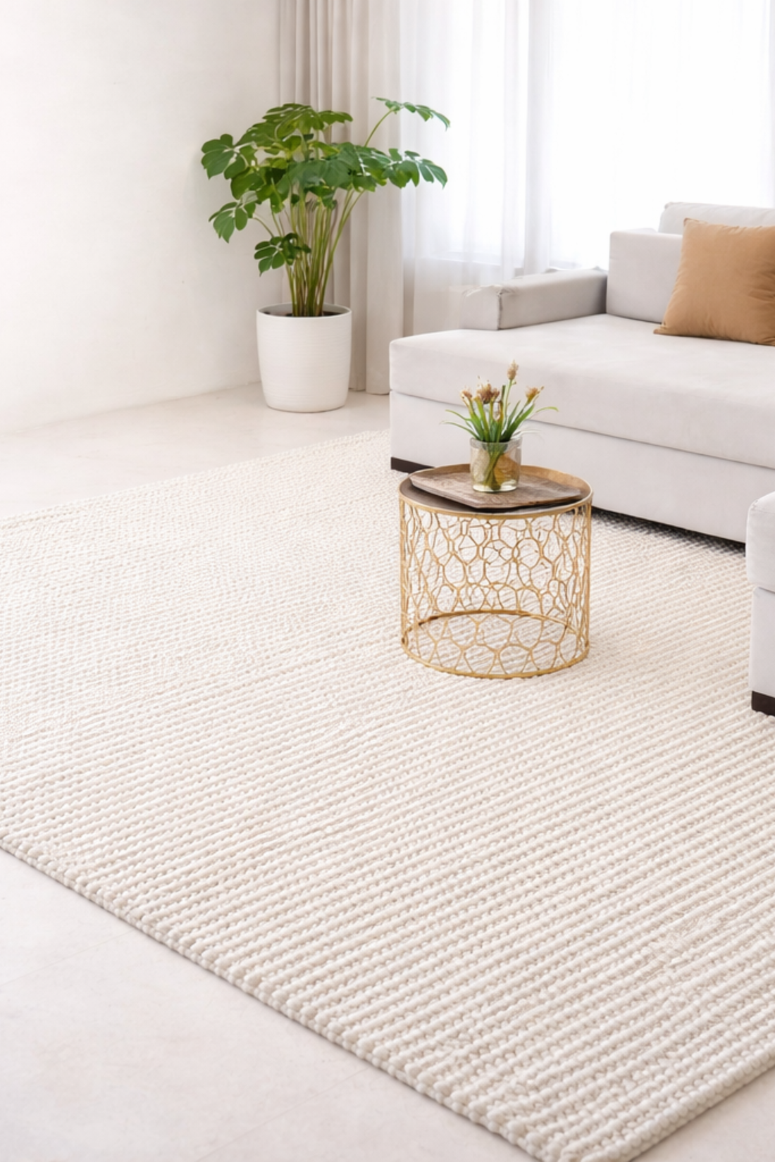 A modern living room with a white textured rug, a light gray sofa with a beige cushion, a gold wire coffee table with flowers, a potted plant, and sheer curtains letting in natural light.