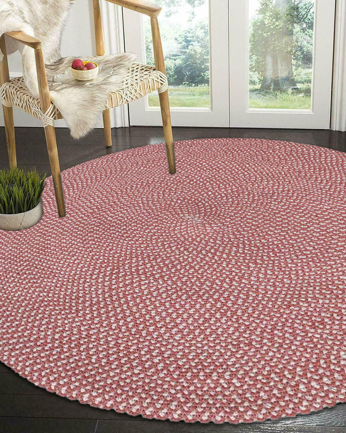 The RAMSHA HOME Handmde Pink Color Round Rug BR 011 adds a pop of color on a dark floor, paired with a modern wooden chair with faux fur, apples in a bowl, a potted plant, and large glass doors revealing green scenery.