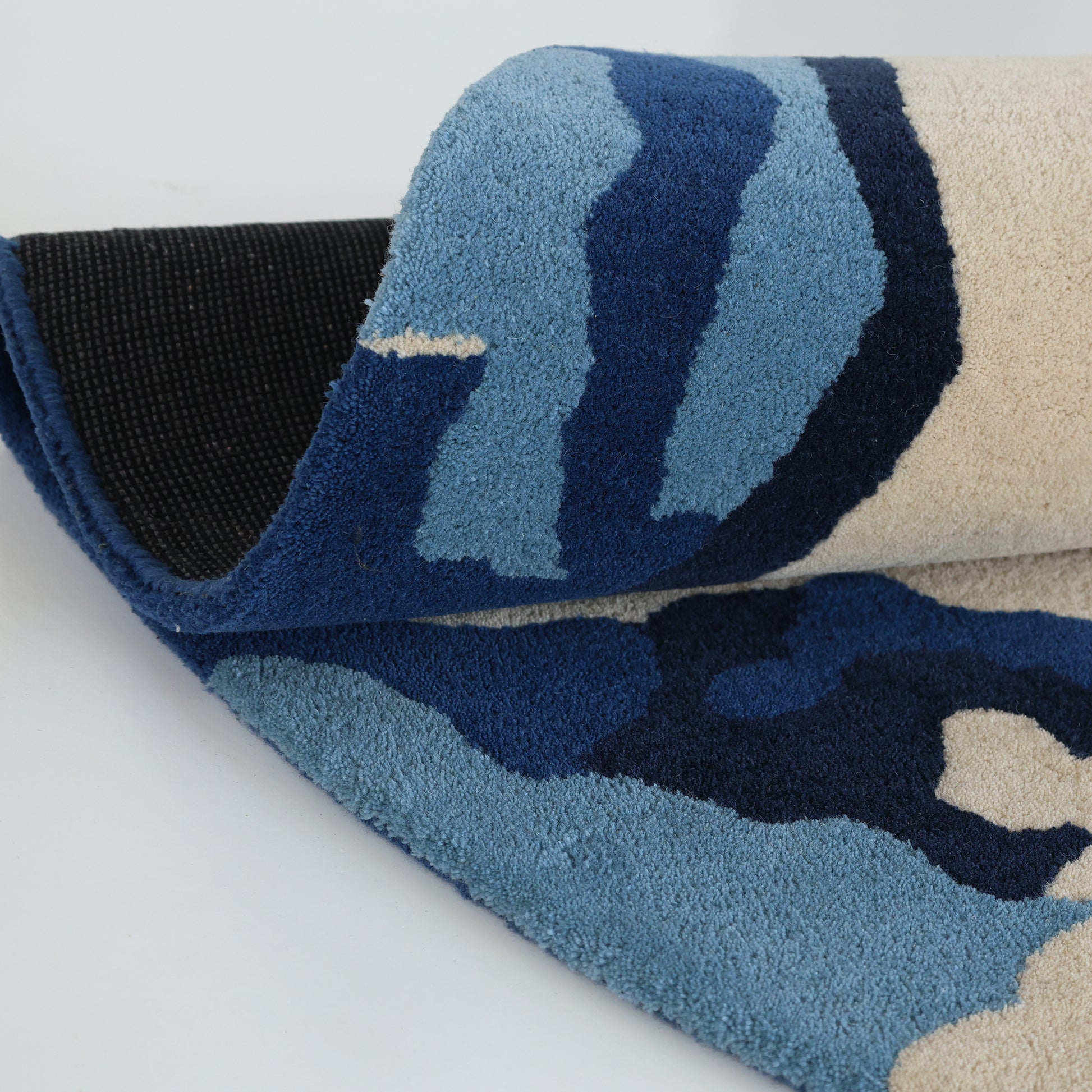 A close-up of the RAMSHA HOME Round Handmade Hand Tufted Carpet HTW-64 showcases its blue, navy, and cream abstract design and black non-slip backing, with the texture of the fibers clearly visible.