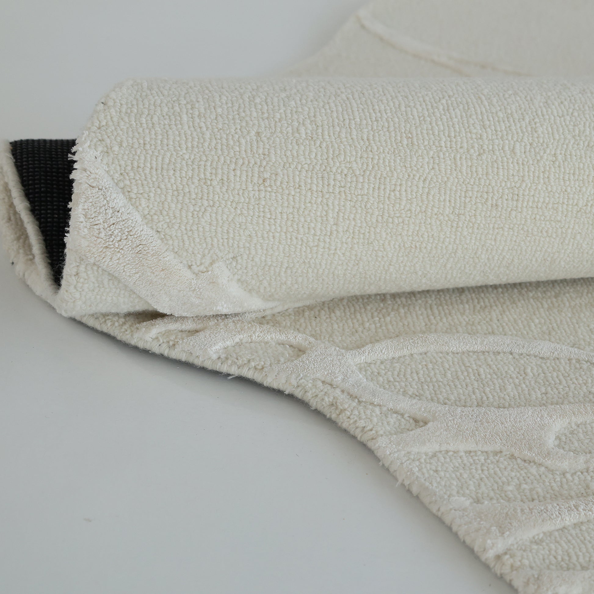 A close-up of the RAMSHA HOME Premium Beige Handmade Carpet HTWV-100 (200×300 cm) shows it partially folded back, revealing a black non-slip underside and subtle raised patterns on its cream-colored wool surface.