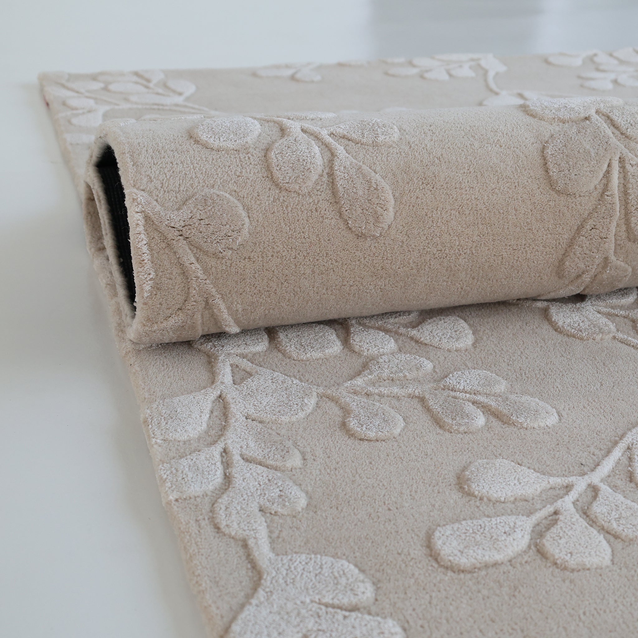 The Artisan Handmade Decorative Area Rug – HTWV-116 by RAMSHA HOME, featuring a beige hand-tufted wool design with a raised leaf pattern, is partially rolled up on a white surface.