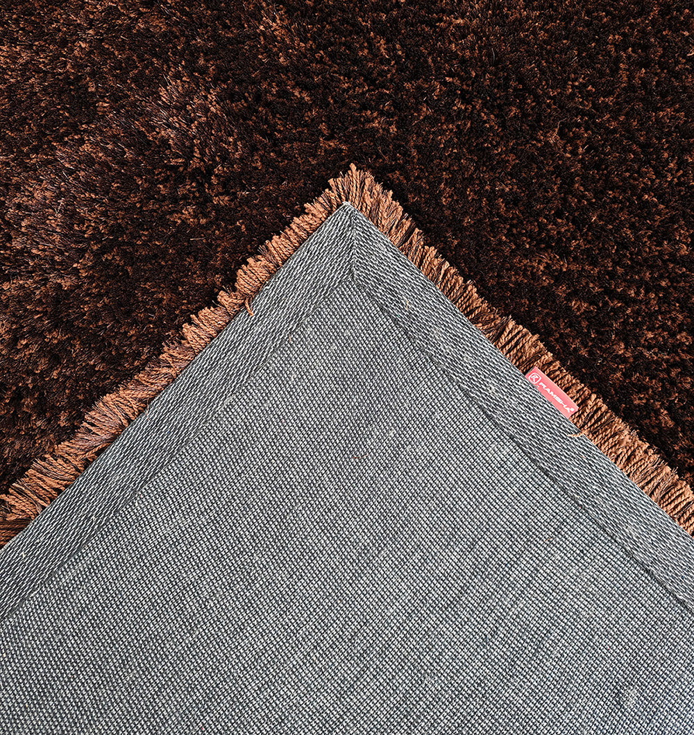 A close-up of the RAMSHA HOME Fluffy Shaggy Area Rug SG 04 in brown, showing its plush texture and rich color where it overlaps with a modern grey rug featuring brown fringe. The focus is on one corner, emphasizing textures and contrast.