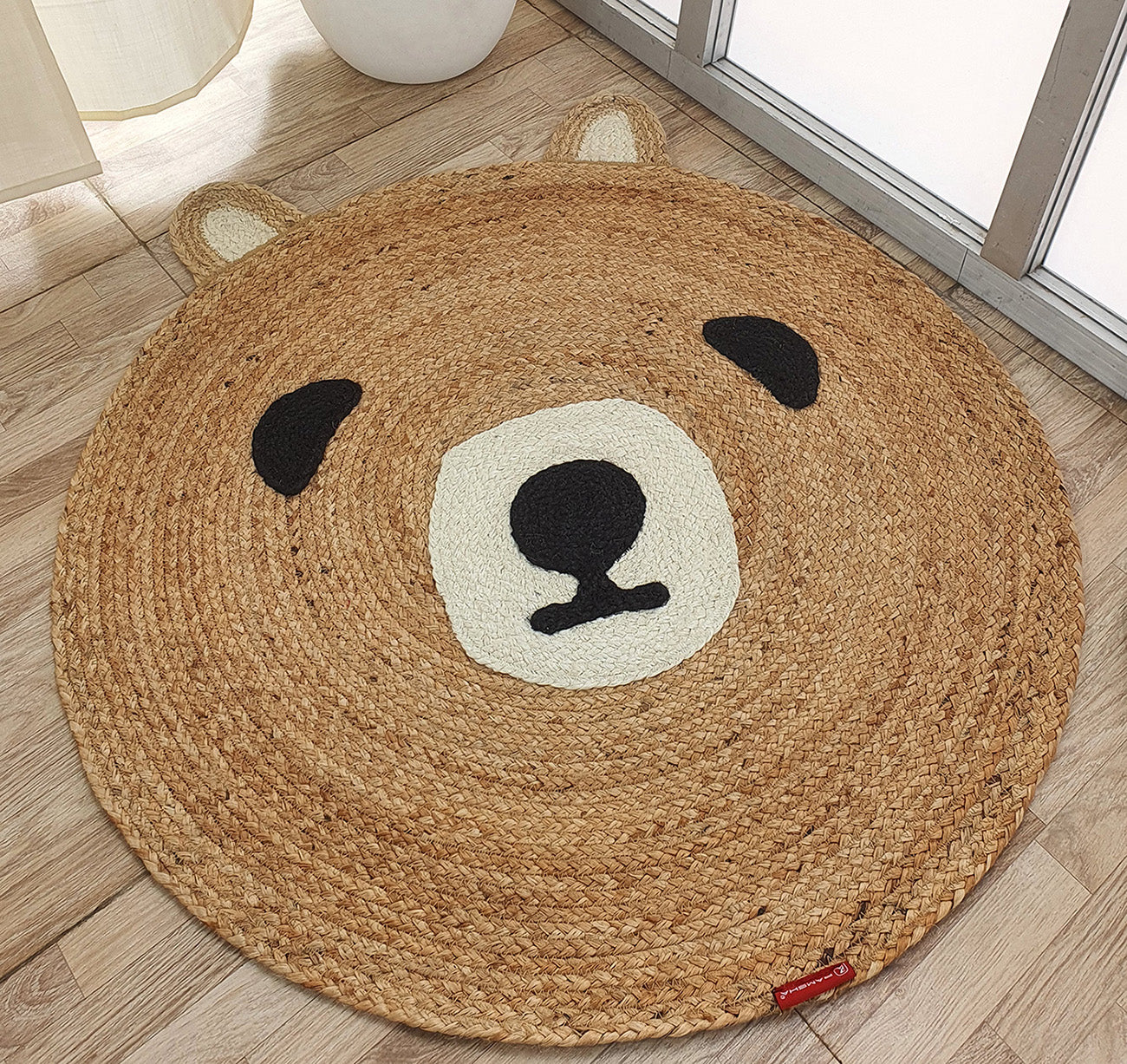 The RAMSHA HOME Amazing Handmade Bear Shape Jute Rug - BR 022 (90cm) features a tan round bear face with beige ears and snout, plus black eyes and nose, shown on a wooden floor near a window.