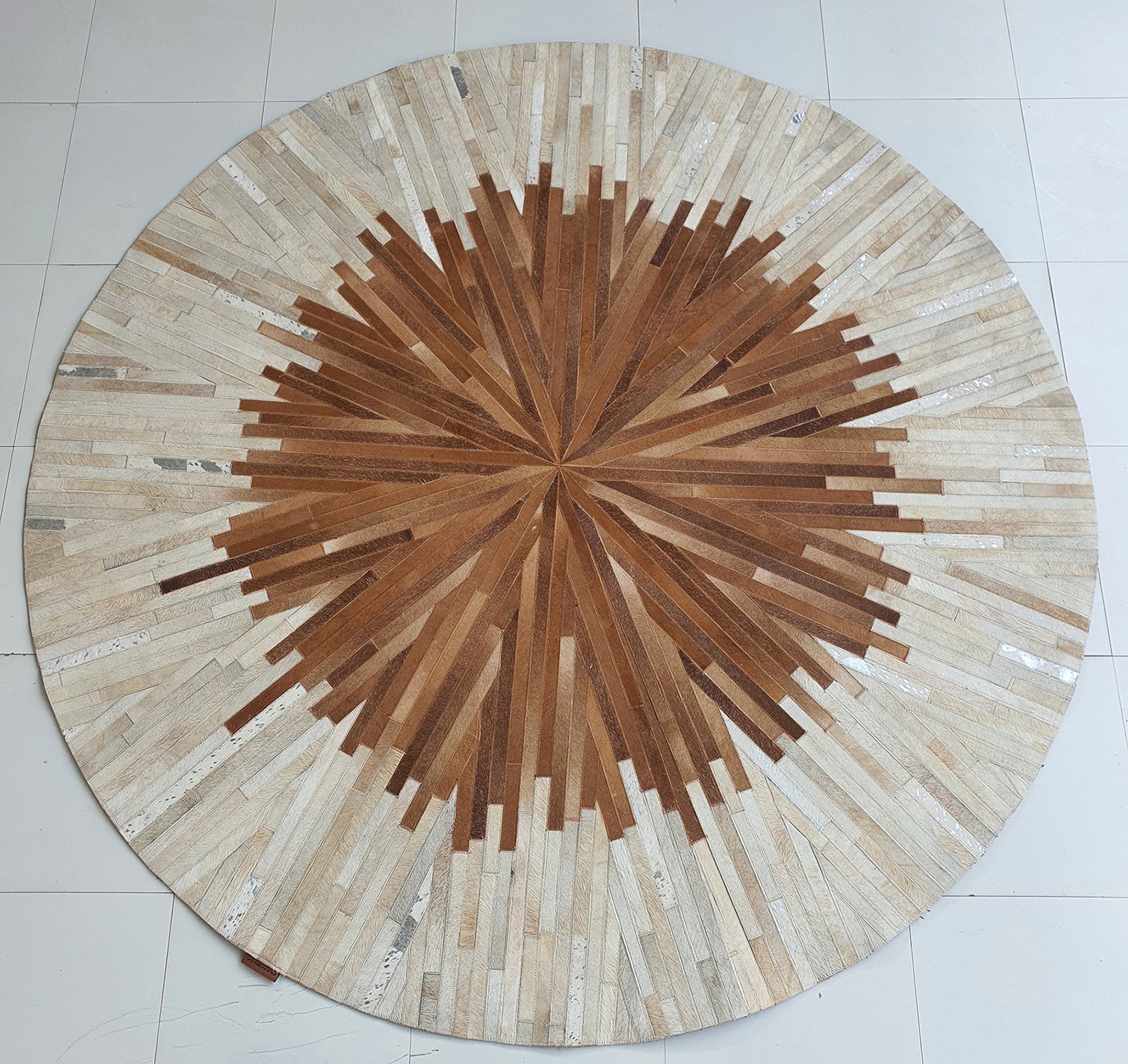 The Multi Color Round Leather Rug - LR 016 features a modern radial pattern in beige, brown, and tan segments for a striking sunburst effect, beautifully displayed on a white tiled floor.