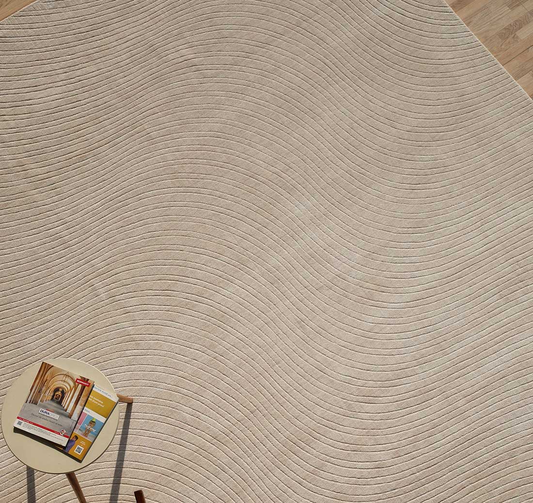 The calm, minimalistic space features the RAMSHA HOME Natural Handmade Area Rug - LKH-03 (300x400 cm) with its beige textured surface and subtle wavy lines, complemented by a side table holding a magazine, book, and cup in the corner.