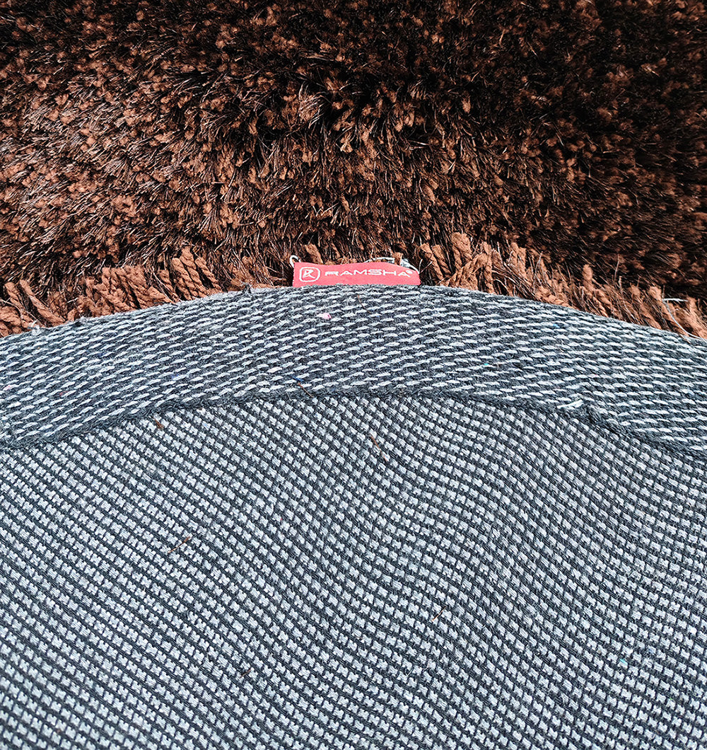 A close-up of the Fluffy Shaggy Round Shape Area Rug SG 04 - Brown by RAMSHA HOME shows its plush brown surface above a textured gray-and-white woven fabric. A small red RAMSHA HOME tag appears near the top center of the image.