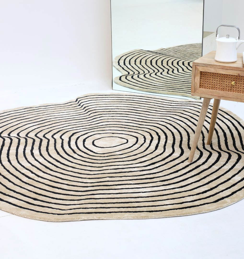 The RAMSHA HOME Round Decorative Area Rug Multi Color HTWV-96, with wavy beige and black concentric lines, is shown on a white floor beside a wooden table and a large mirror reflecting its distinctive pattern.