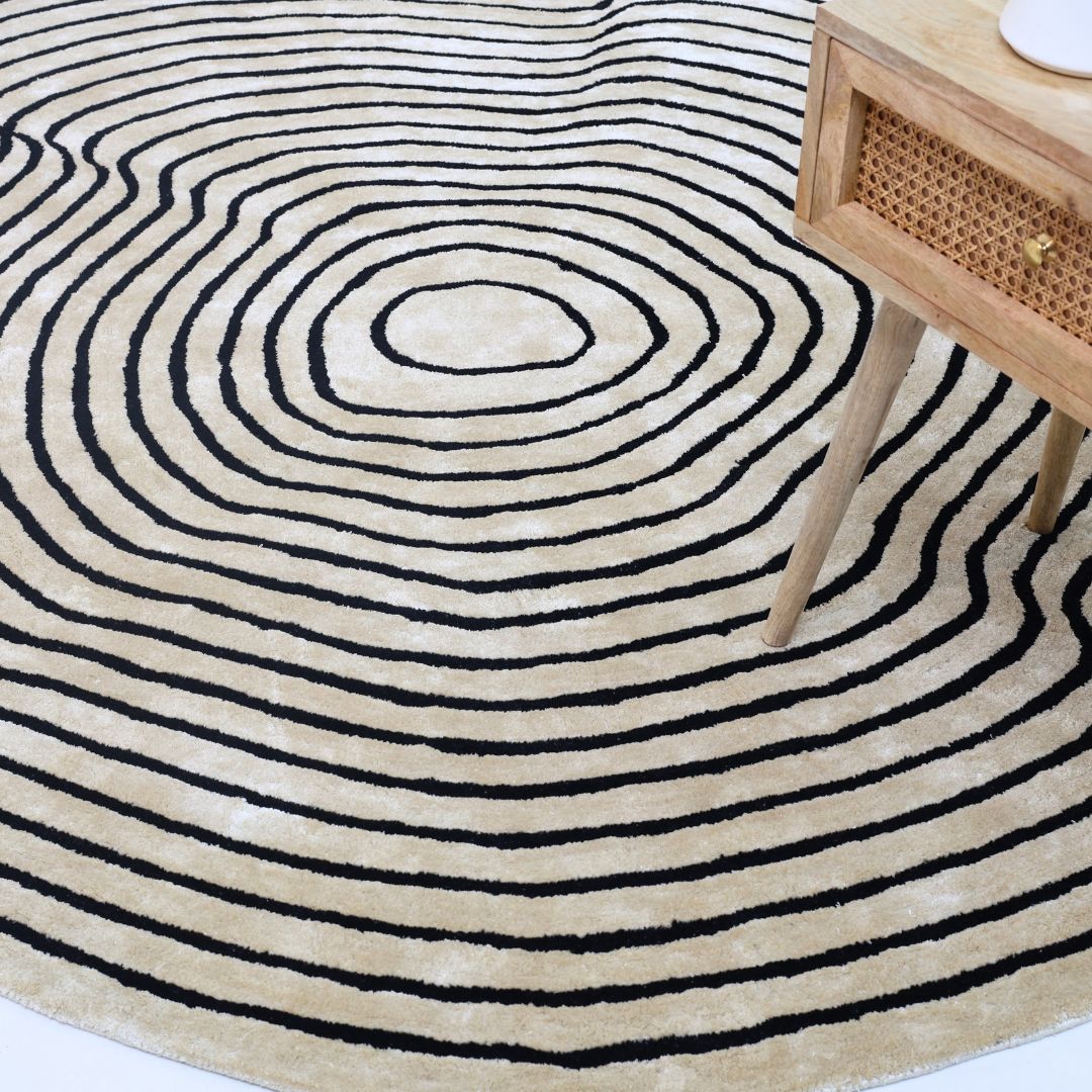 The RAMSHA HOME Round Decorative Area Rug Multi Color HTWV-96, featuring concentric black rings, is partially visible beside the wooden leg of a small table with a woven panel and a round white object on top.