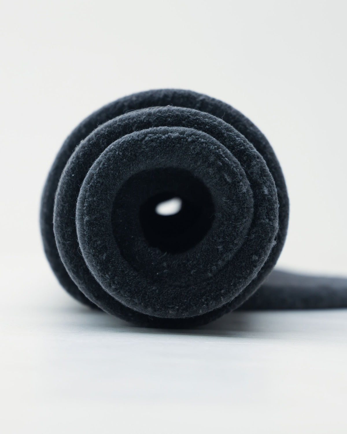 A close-up of a rolled-up black yoga mat with a soft, textured surface, viewed from the end. The mat rests on the RAMSHA HOME Oval Shape Home Decor Multi Color Rug HTWV-90, which is placed against a plain background.