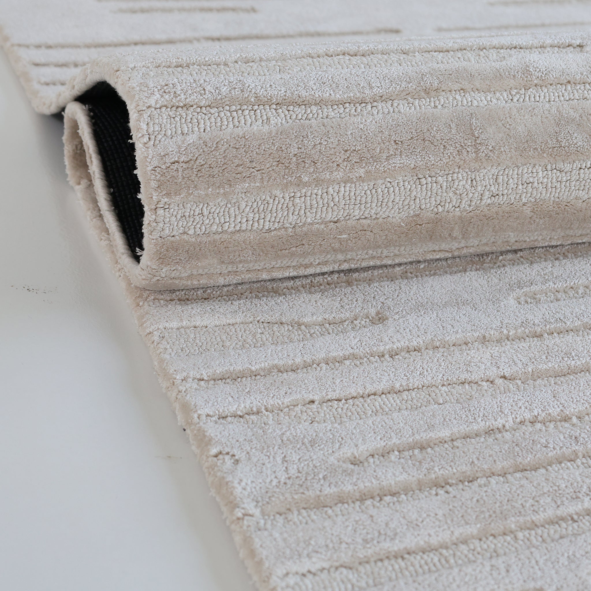 The RAMSHA HOME Home Decor Handmade Area Rug - HTV 17 features a neutral textured design with subtle stripes, hand-tufted for understated elegance—partially rolled on a smooth white surface, perfect for any room.