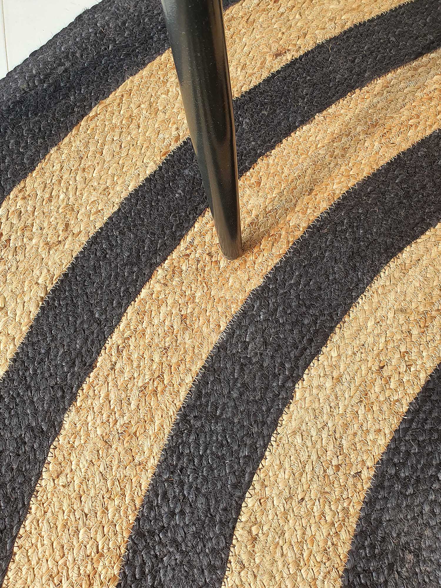 Close-up of the RAMSHA HOME Handmade Braided Jute Floor Round Rug - BR 058 (200cm), featuring alternating tan and black curved stripes, partially covered by a black furniture leg.