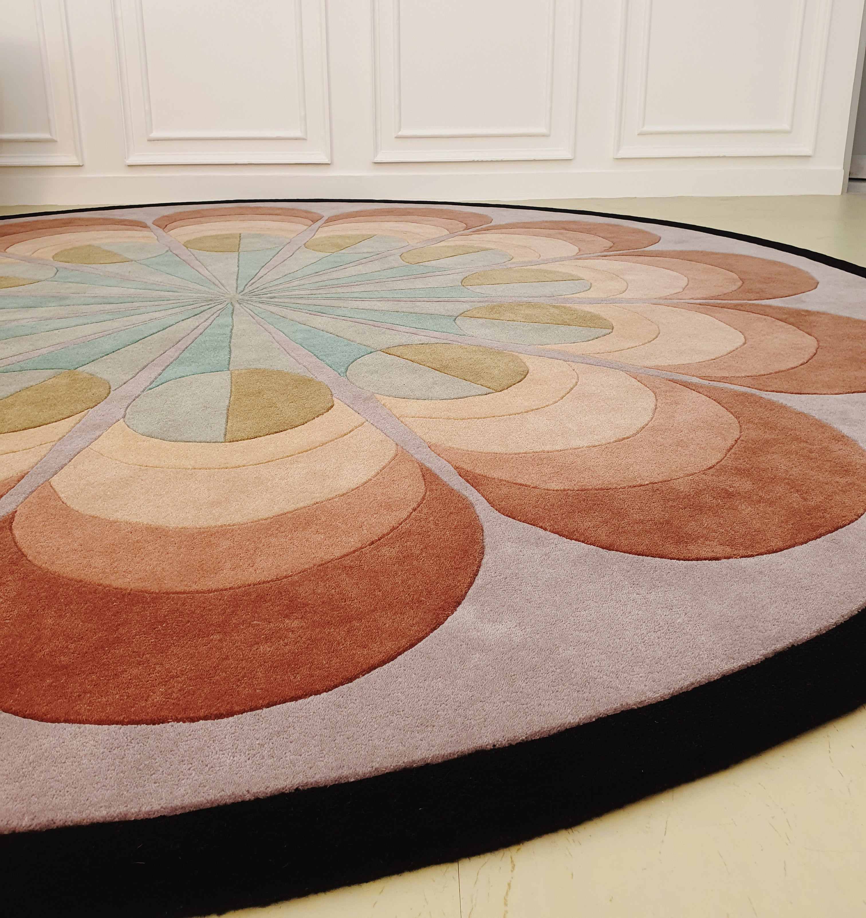 The RAMSHA HOME Handmade Carpet for Bedroom Decor – HTW-54 features a round wool design with a geometric radial pattern in beige, brown, peach, and light green, black-edged and perfect for adding modern style and warmth to any space.