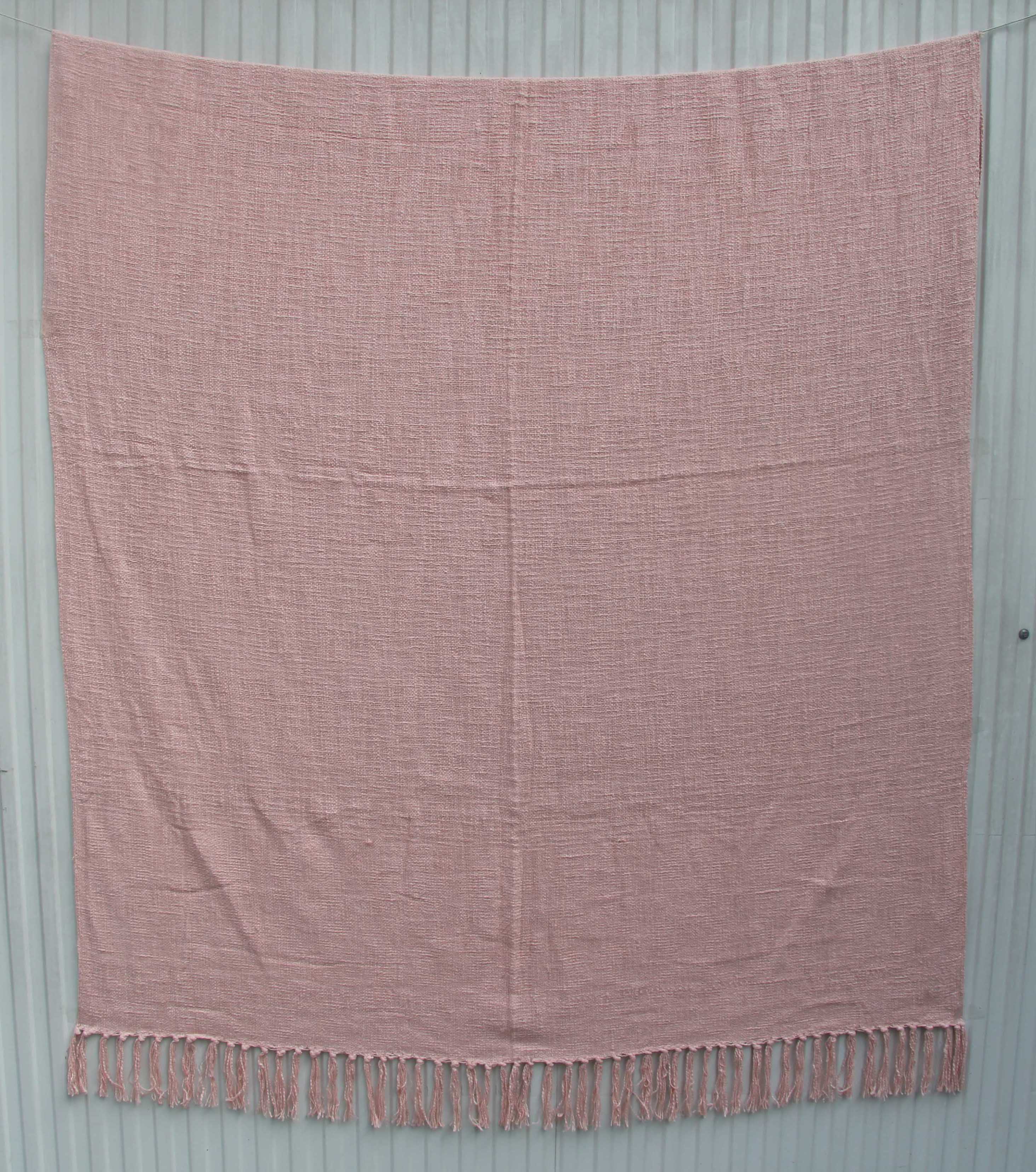 The Pink Color Cotton Throws for Sofa TAL 042 (125x150 cm) by Ramsha Home offer breathable comfort and a textured feel, with fringed tassels, elegantly displayed against a light gray wooden background.