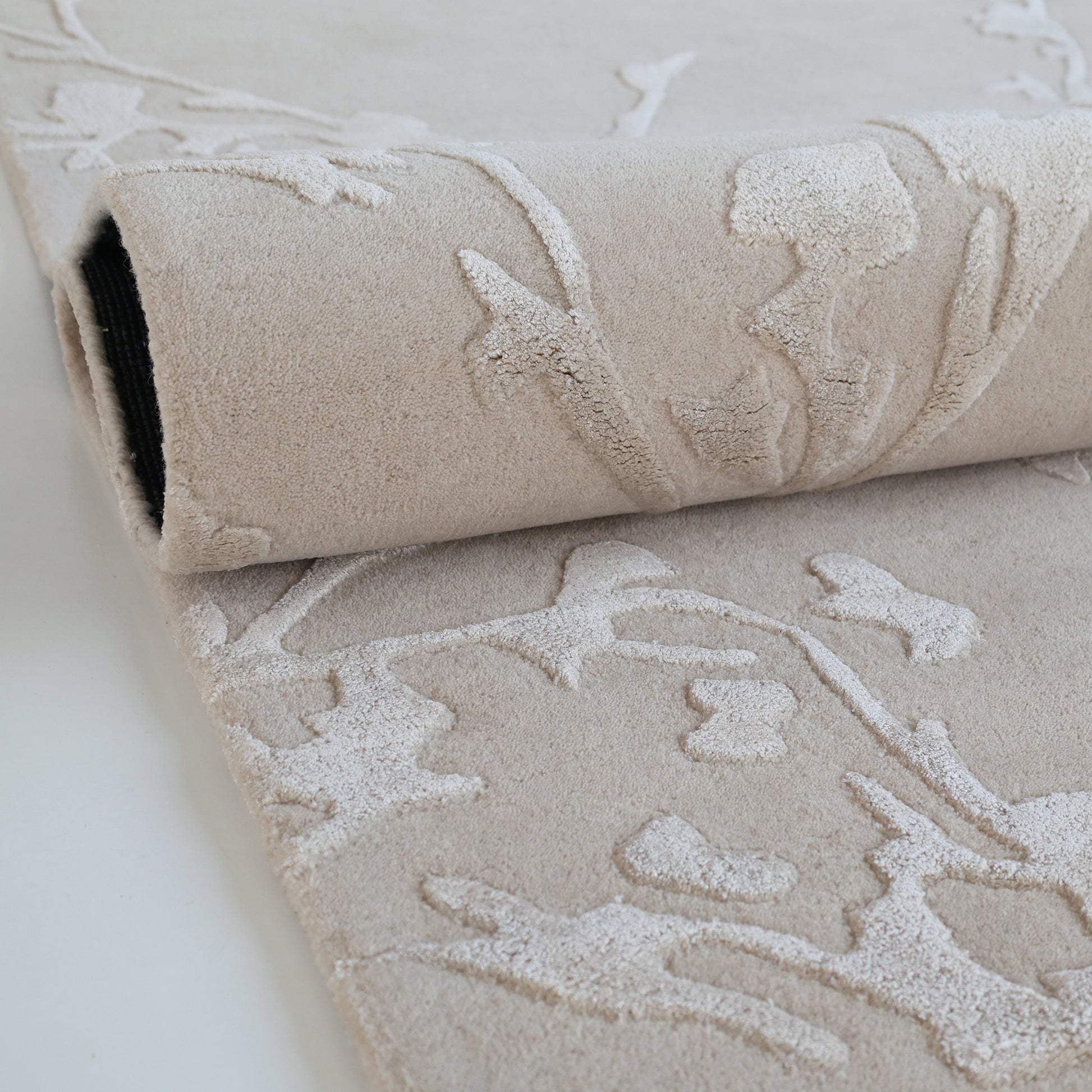 A close-up of RAMSHA HOME's Handmade beige Color Decor Rug- HTWV 115, partially rolled. This hand-tufted wool rug has a raised floral pattern for textured contrast, making it a stylish neutral anchor for any room.