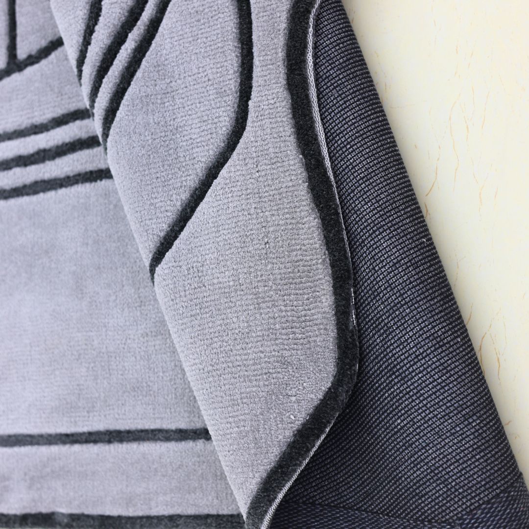 A close-up of the RAMSHA HOME Shaped Rug Grey & Black Color Carpet (CMN-05) shows black line patterns on a grey surface, with a folded corner revealing its textured black underside and unique edge, highlighting its irregular design.