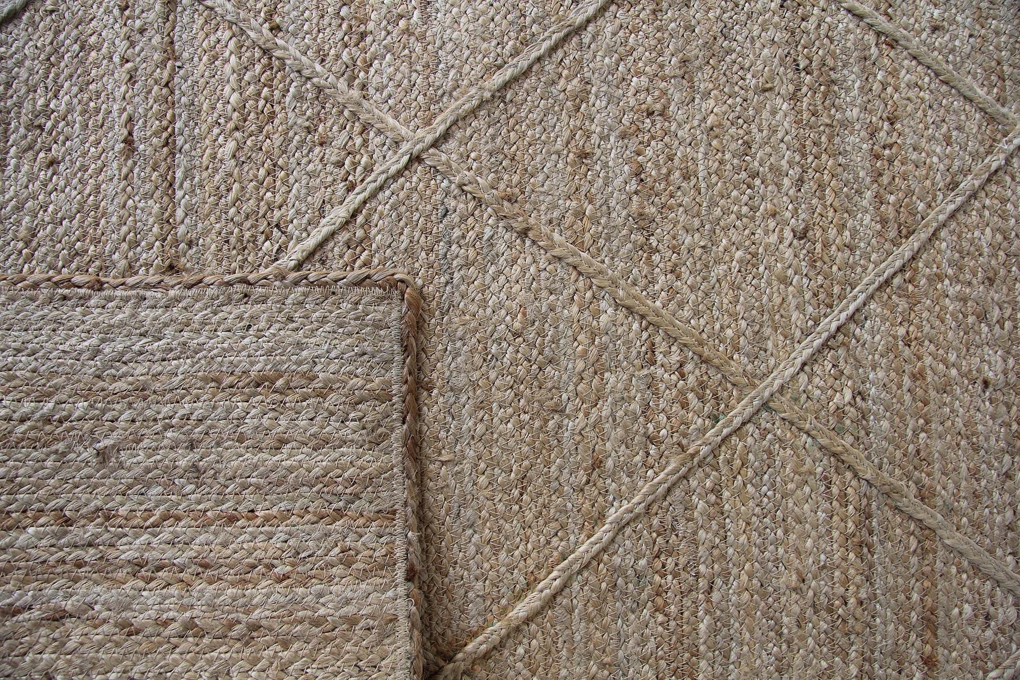Close-up of the Natural Handmade Jute Braided Rug - BR 073 by RAMSHA HOME, featuring chevron and diamond patterns in beige and tan. A smaller rectangular rug overlaps one corner, showcasing a similar braided texture.