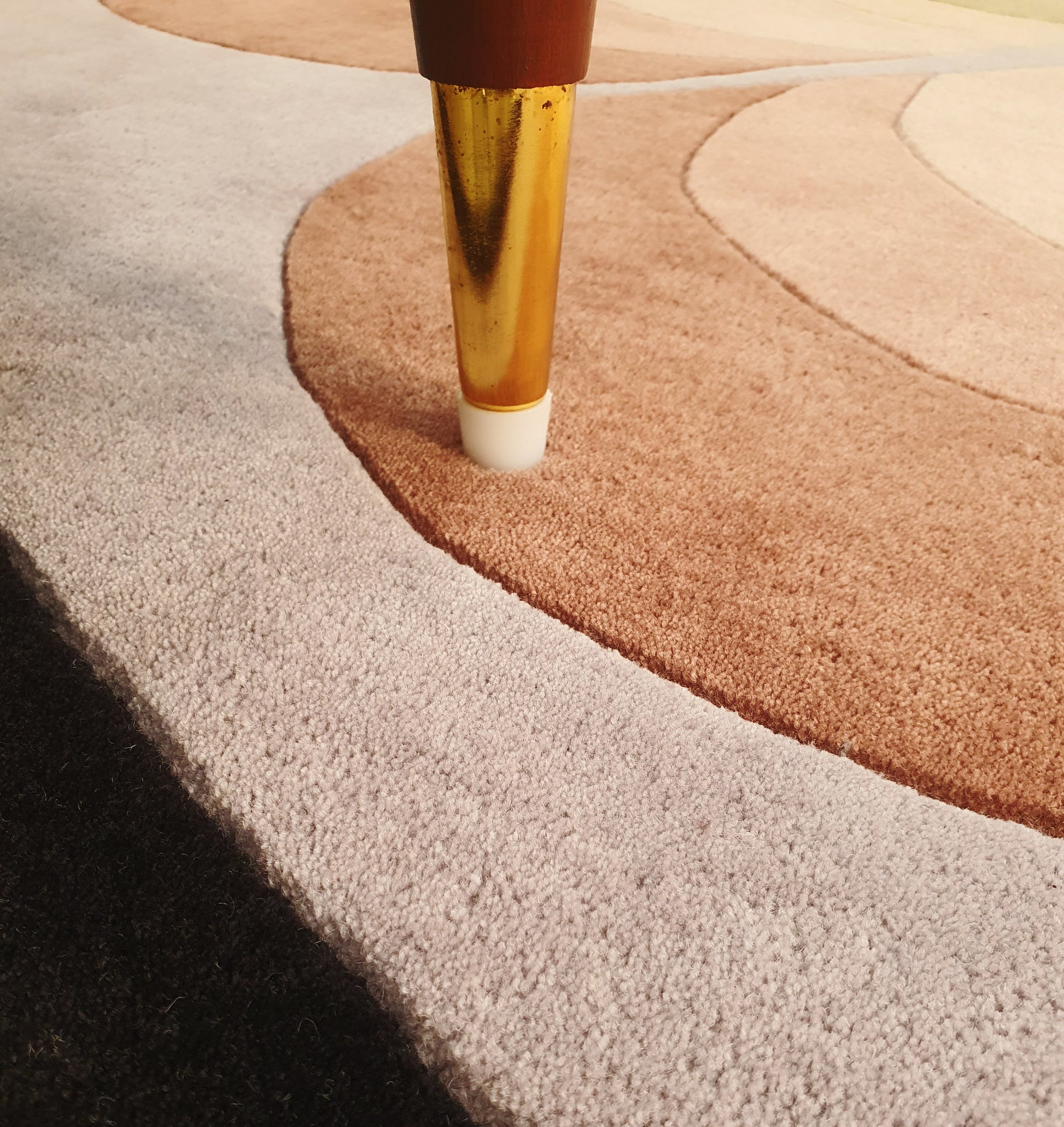 Close-up of a golden furniture leg with a white tip on the RAMSHA HOME Handmade Carpet for Bedroom Decor – HTW-54, a hand-tufted rug with curved patterns in beige, brown, and gray.