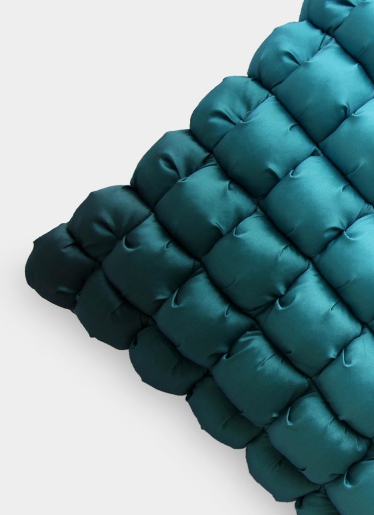 A close-up of the RAMSHA HOME Polyester Bubble Cushion Cover shows its teal, quilted, puffy texture and smooth, shiny fabric against a plain white background with only part of the cushion visible.