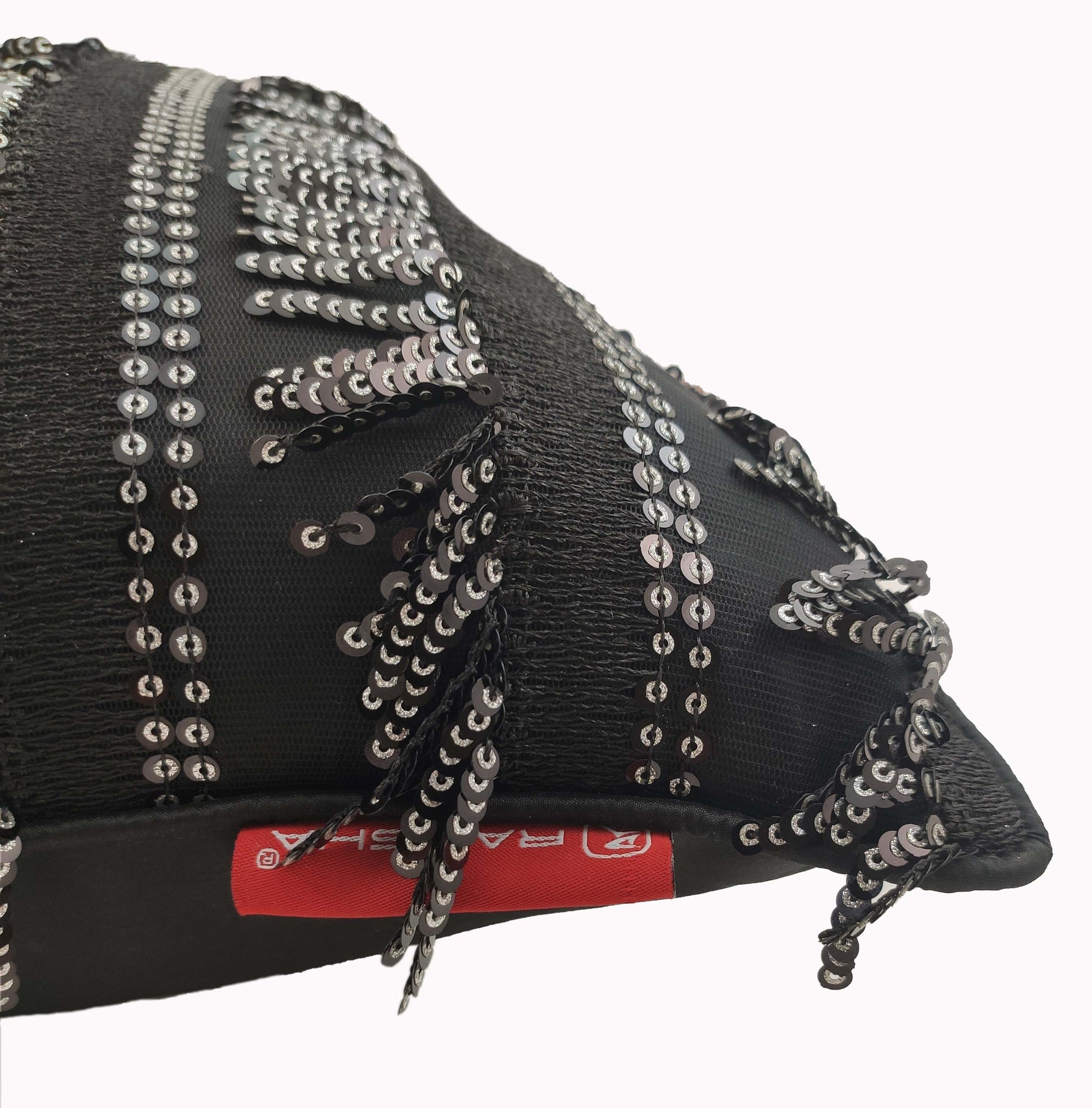 A close-up of the RAMSHA HOME Modern Polyester Cushion Cover in black, decorated with shiny silver sequins and fringe, and featuring a visible red label with white text on the side.