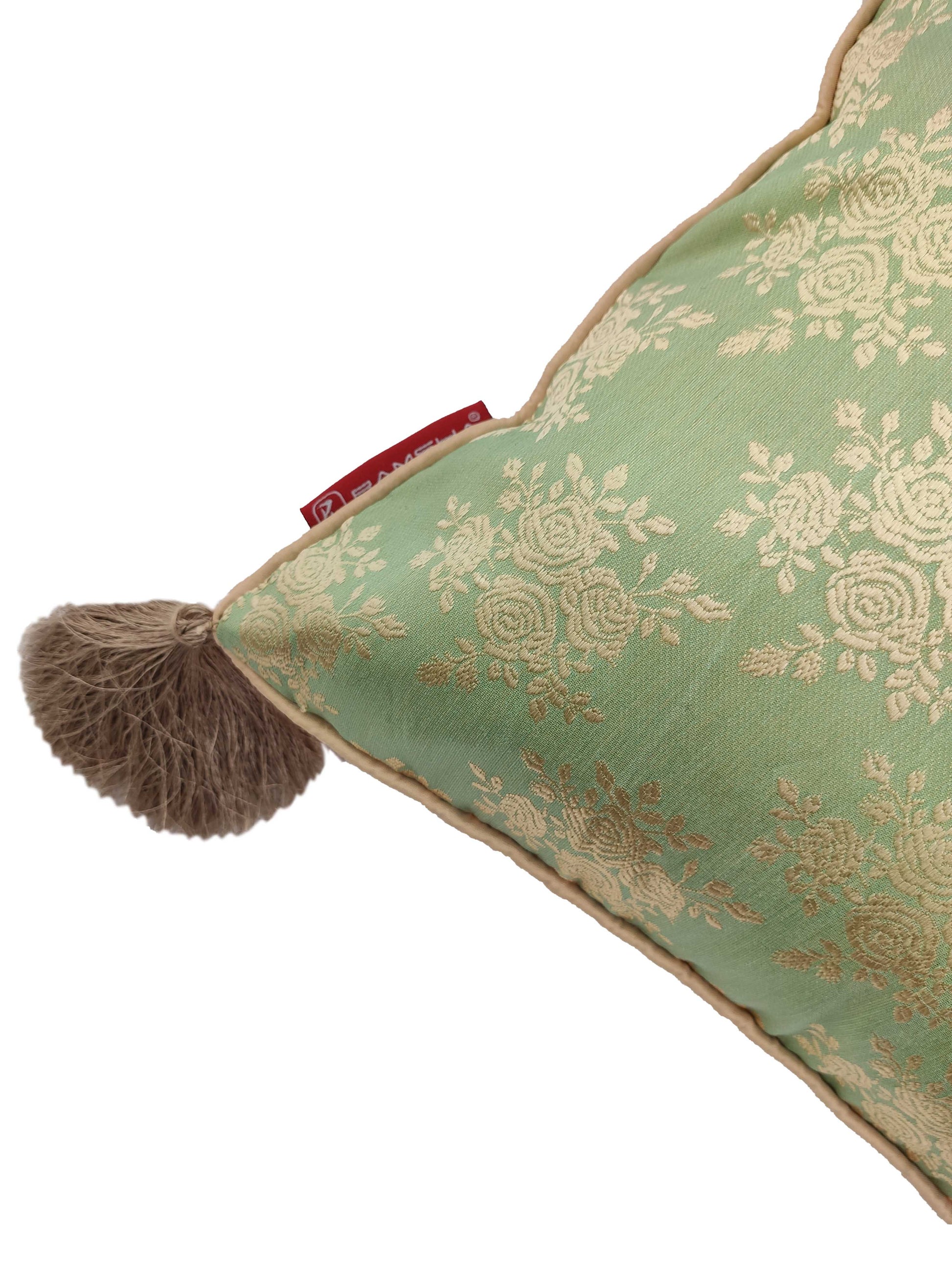 A close-up of RAMSHA HOME’s Polyester Cushion Cover With Tassel CS-042 shows green fabric with gold floral patterns, a beige braided trim, a large brown tassel on the corner, and a small red fabric tag near the tassel.