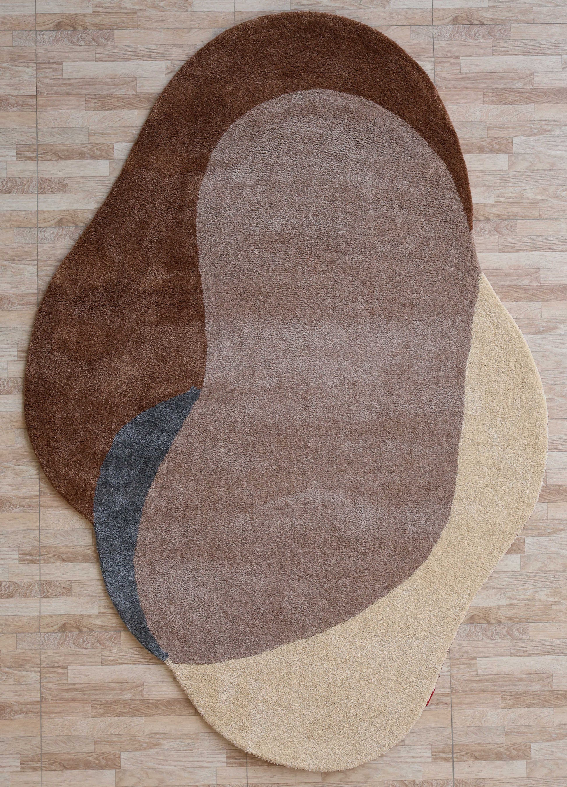 The RAMSHA HOME Contemporary Freeform Plush Rug - SG 047 features overlapping bean-like patches in brown, beige, gray, and tan on a light wood-patterned floor, creating a modern abstract look with an irregular shape.