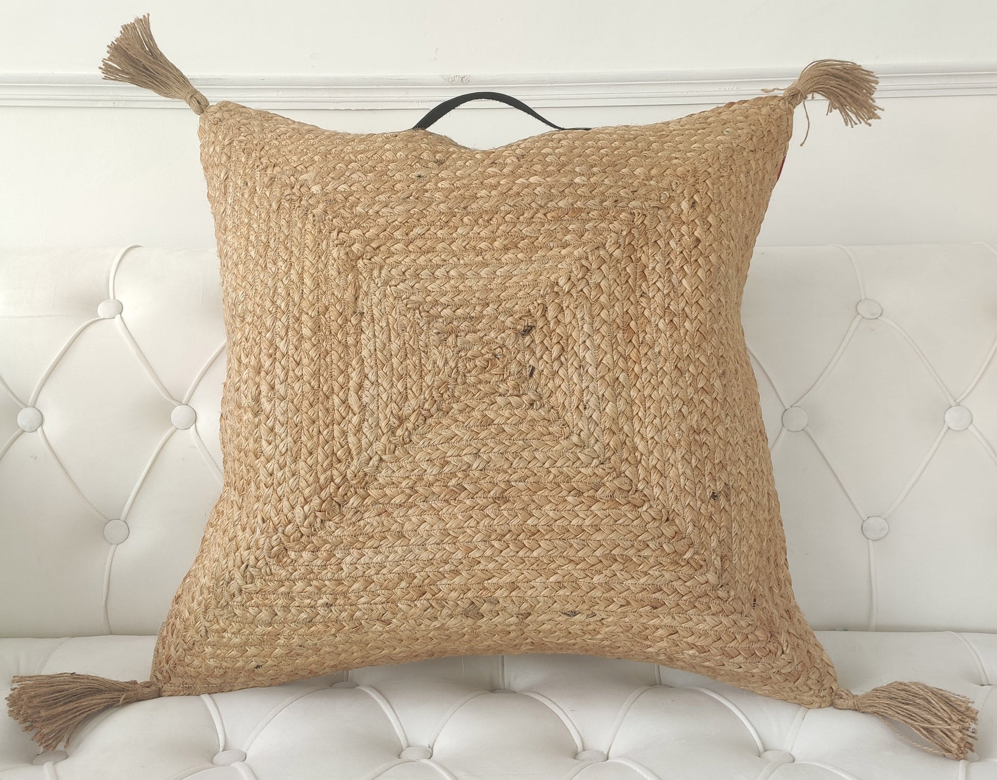 The RAMSHA HOME Jute Cotton Cushion With Tassel And Handle CS-195, featuring a braided texture and corner tassels, is displayed on a white, tufted sofa.
