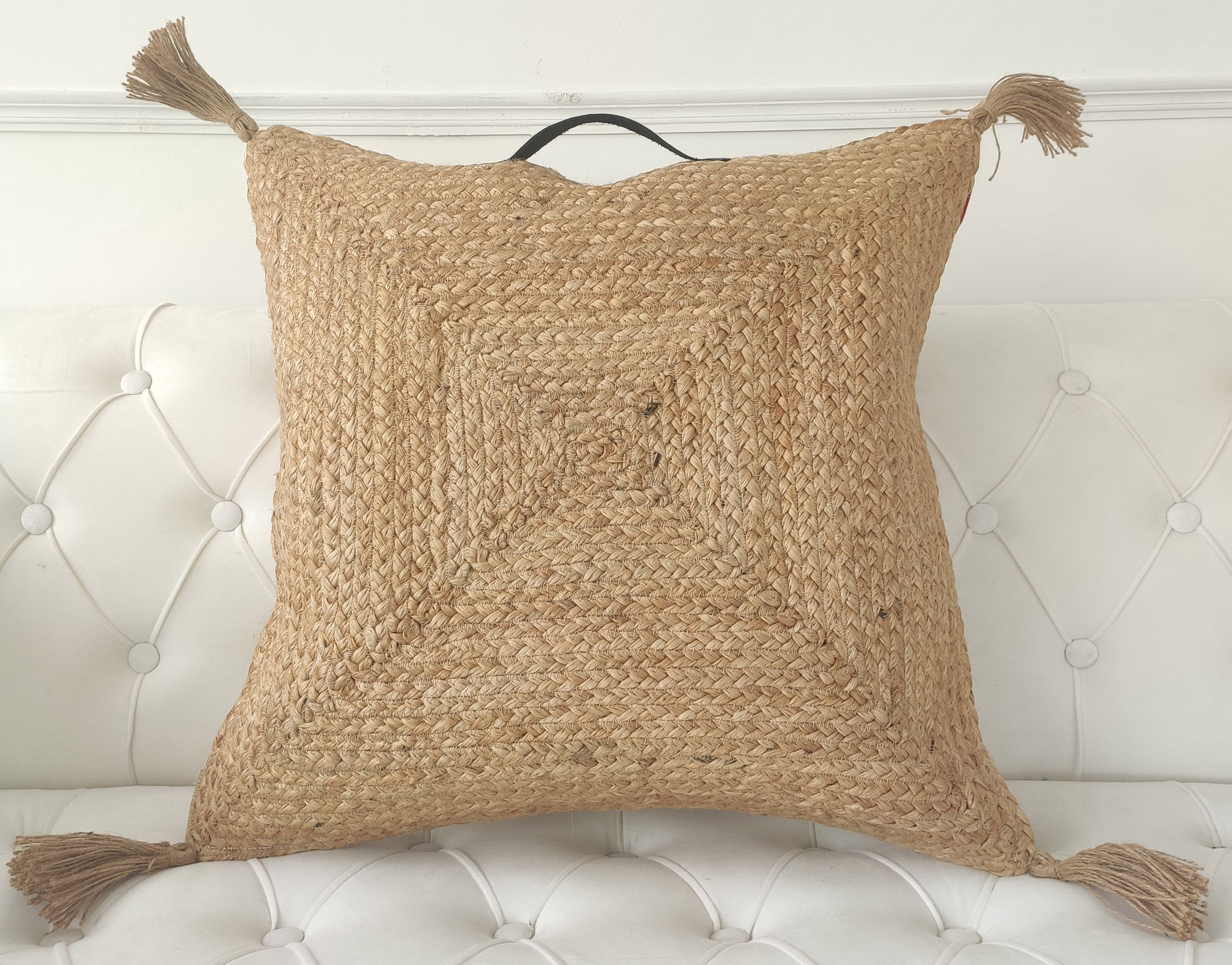 The RAMSHA HOME Jute Cotton Cushion With Tassel And Handle CS-195, featuring a braided texture and corner tassels, is displayed on a white, tufted sofa.