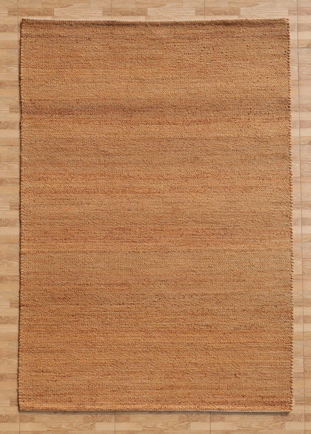 A rectangular Handmade Natural Color Hand Woven Area Rug SR-064 by RAMSHA HOME, with its natural brown jute fibers and subtle linear texture, lies on a light wood floor. This artisanal area rug features handmade craftsmanship and gentle color variations, bringing a simple and earthy elegance that enriches any home décor.