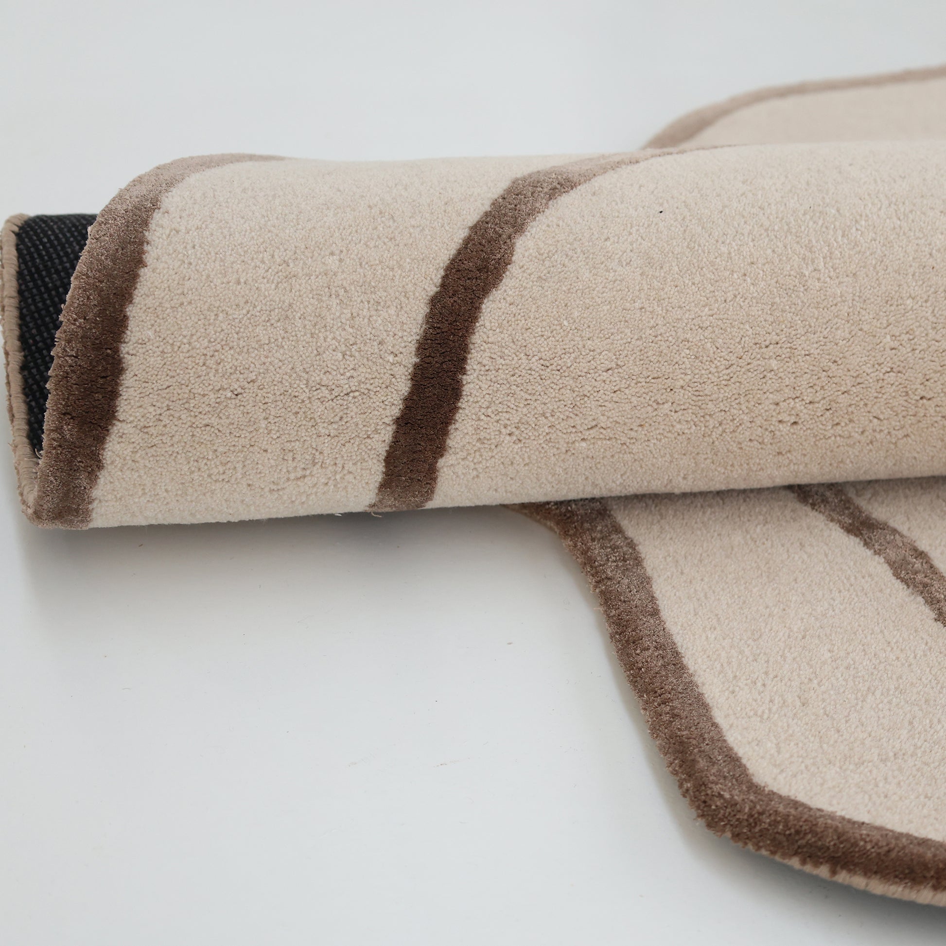 A close-up of the RAMSHA HOME Handmade Multi Color Hand-Tufted Area Rug HTWV-65, showing its plush texture and brown stripes, partially rolled on a white surface with dark backing visible at the edge.