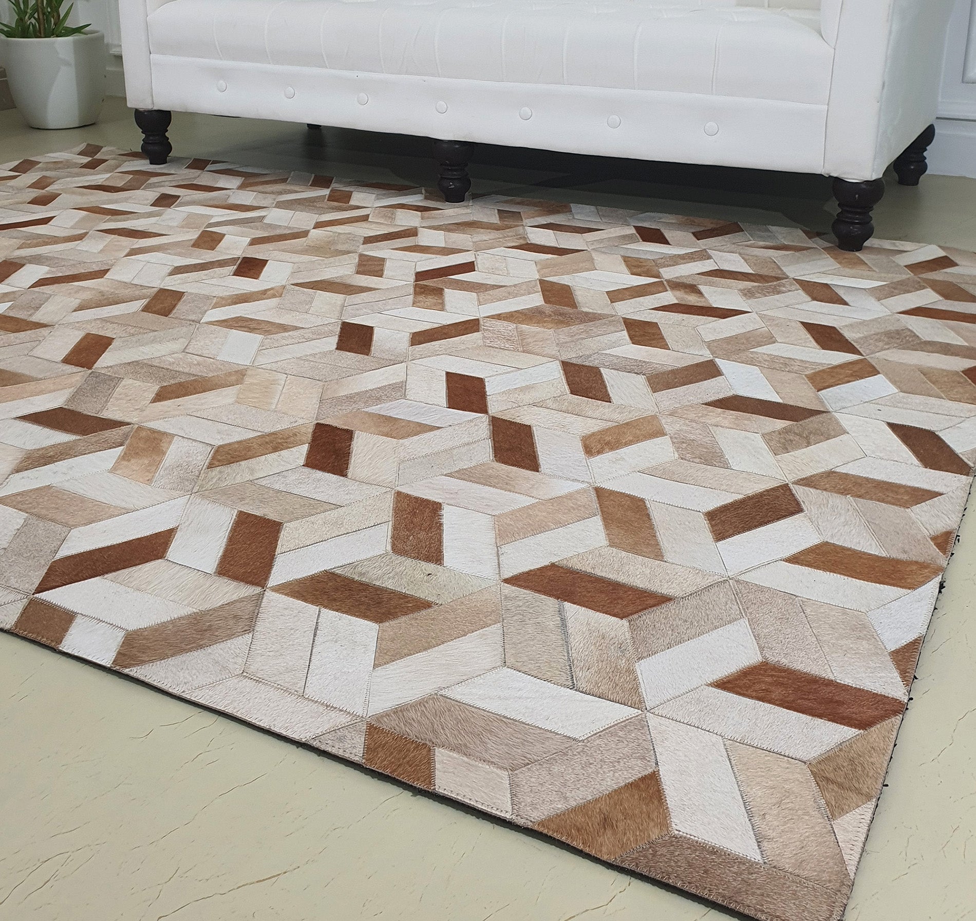The RAMSHA HOME Geometric Cube Pattern Handmade Leather Area Rug LR-012, featuring beige, brown, and cream 3D cubes, is displayed on a light floor before a white sofa, with a small plant visible in the background.