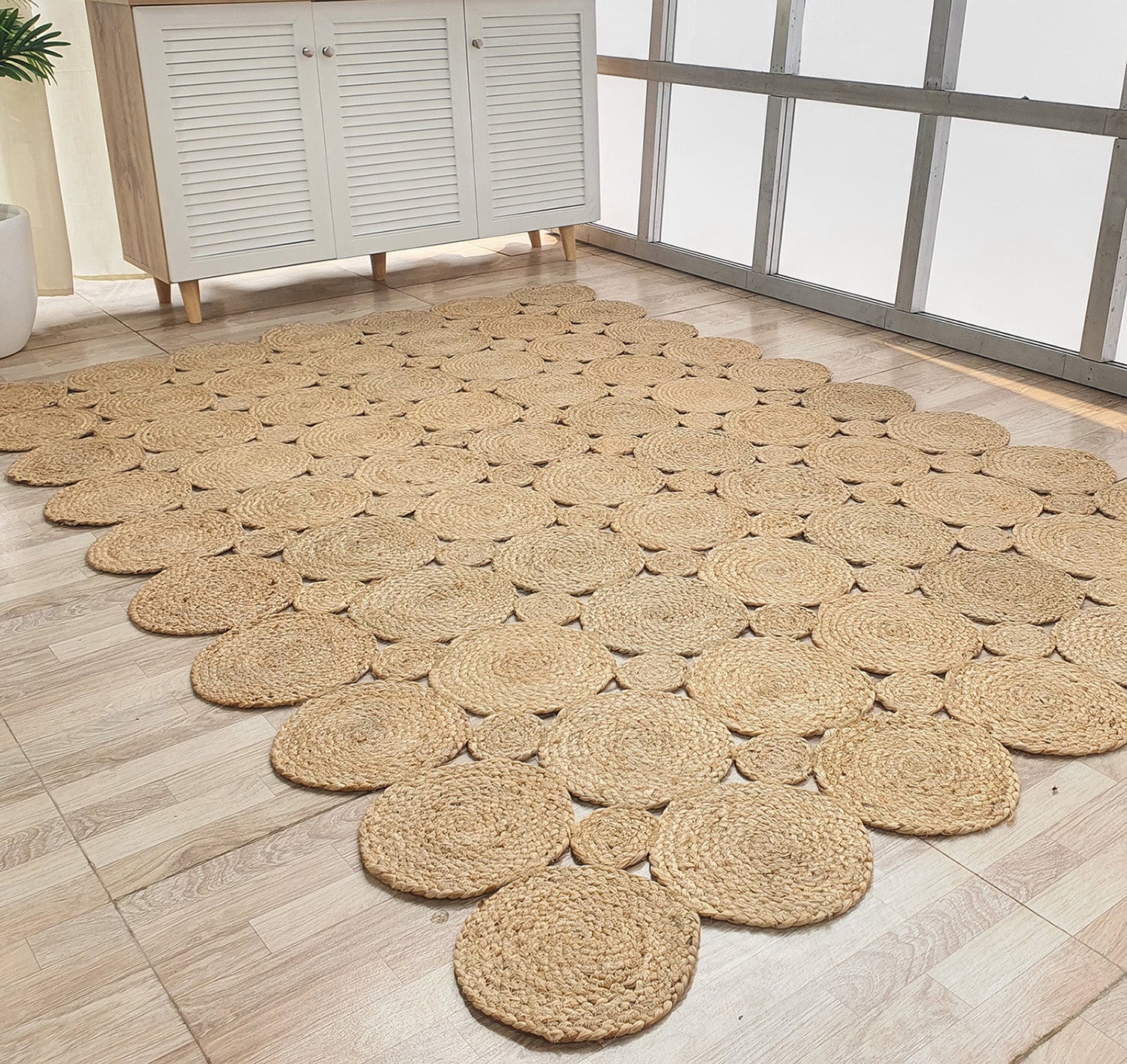 The RAMSHA HOME Handmade Circular Pattern Jute Rug BR-035, featuring round natural fiber circles in a scalloped design, is placed on a light wood floor near a white cabinet and large frosted windows.