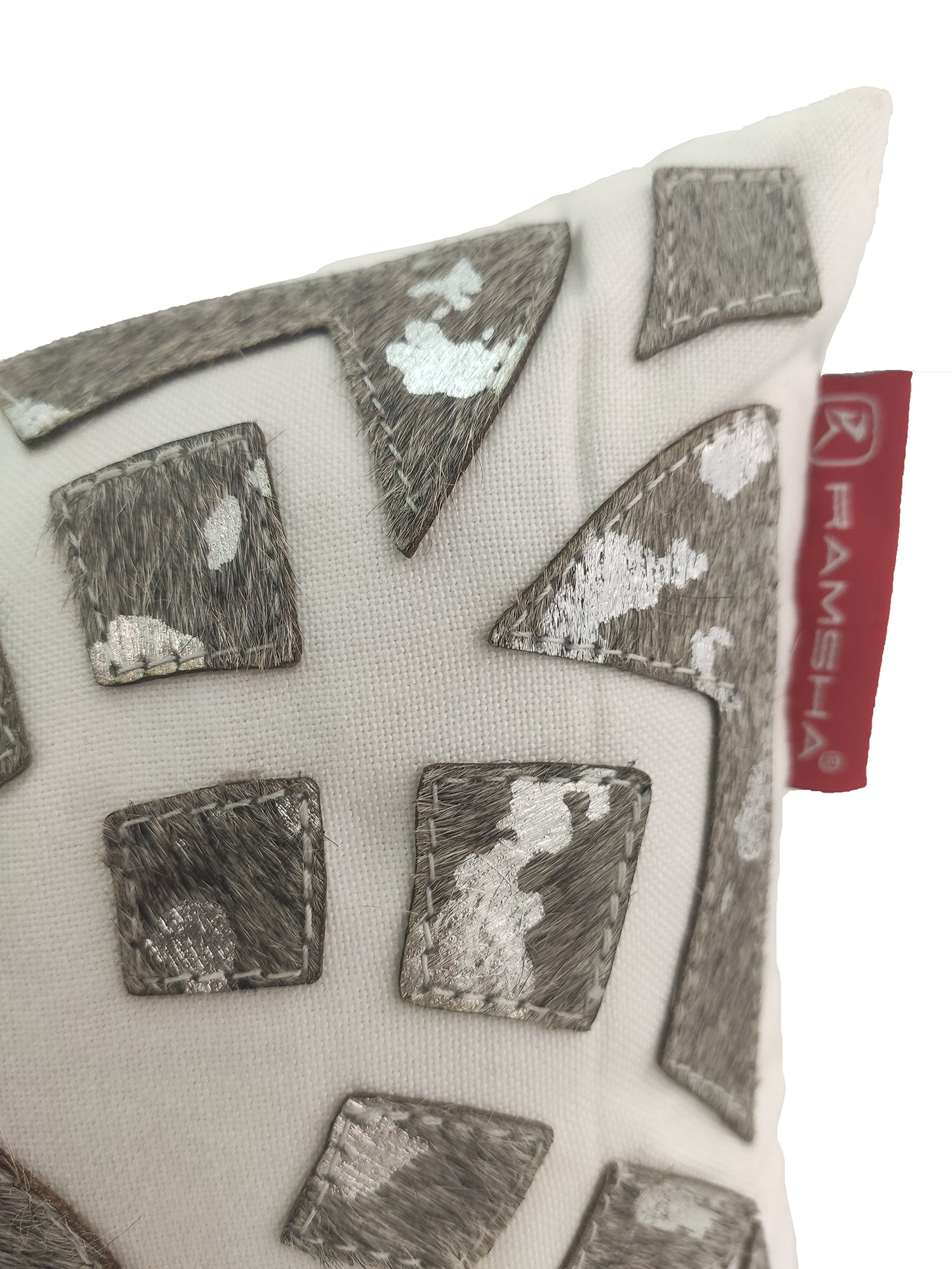 A close-up of the Decorative Cushion CS-105 by RAMSHA HOME, showcasing a white pillow with textured brown and silver geometric patches and a red fabric tag featuring the brand name on the side.