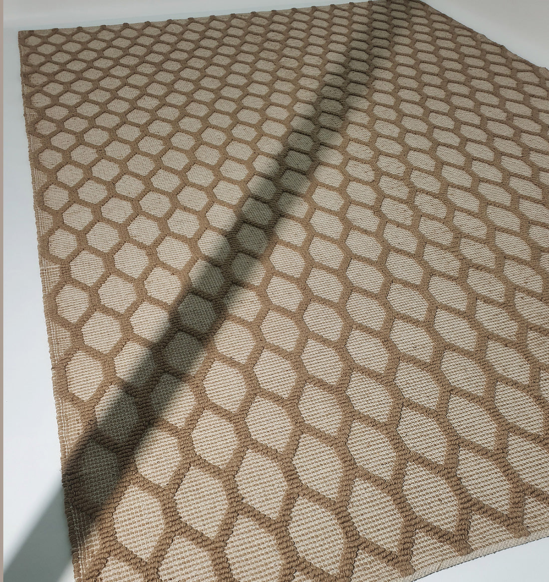 The RAMSHA HOME Rectangle Woven Area Rug Neutral Beige And Brown SR-023, featuring a hexagonal pattern in beige and brown, lies on a white surface with one corner lifted, casting a shadow and gentle wave.