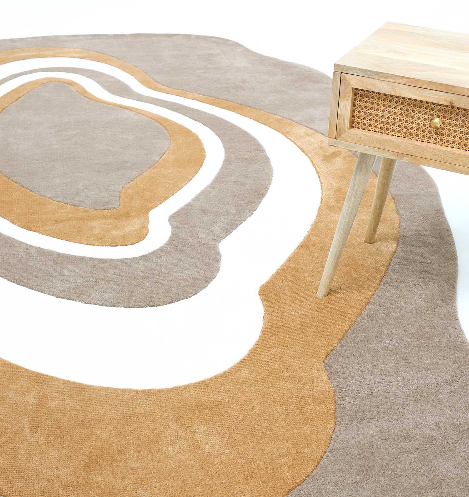 A close-up of the RAMSHA HOME Oval Shaped Novara Multi Colored Carpet 200x300 cm (CMN-06) with wavy brown, tan, and white patterns beside a light wood side table featuring angled legs and a woven drawer front.