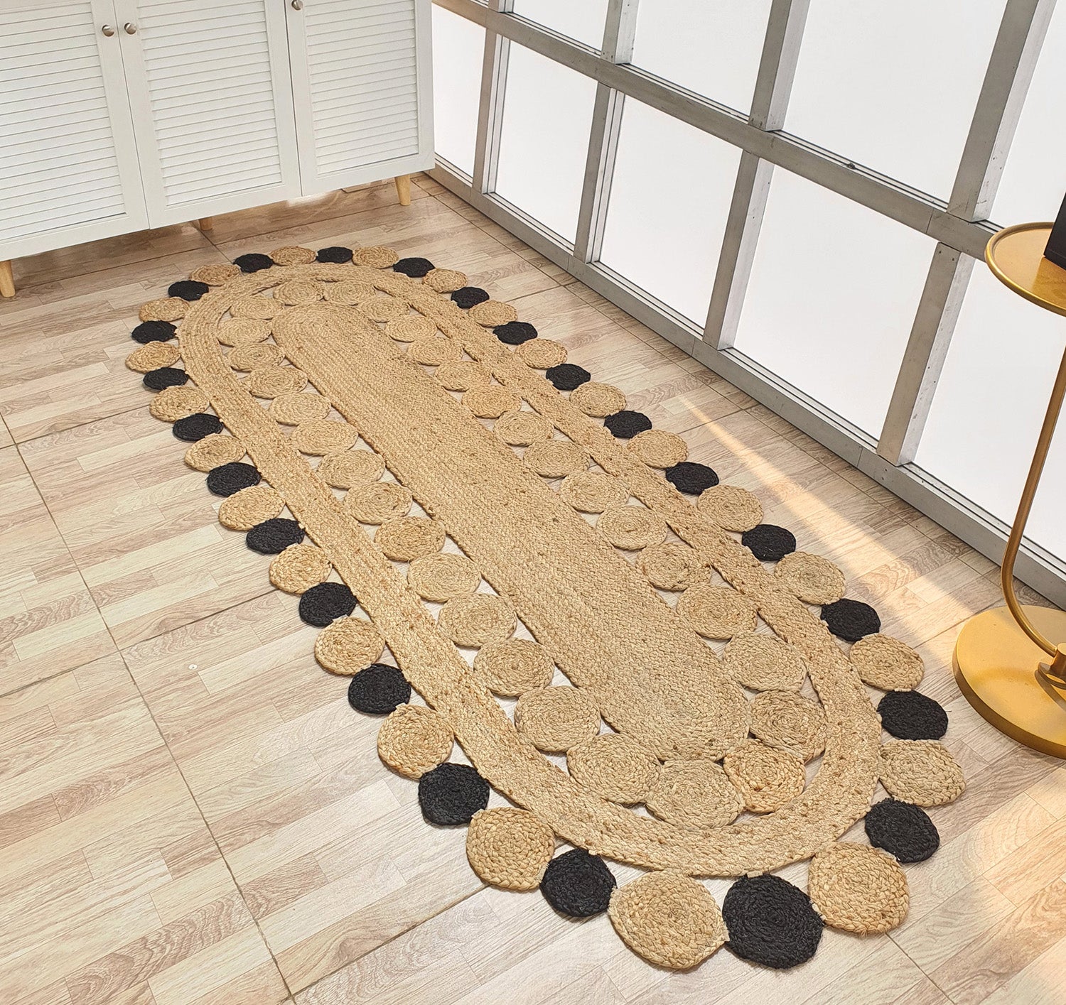 The RAMSHA HOME Oval Shaped Jute Braided Rug Black & Natural BR-033 features black and natural circular patterns and is placed on a light wooden floor by a white cabinet and large metal-framed windows.