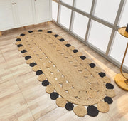 The RAMSHA HOME Oval Shaped Jute Braided Rug Black & Natural BR-033 features black and natural circular patterns and is placed on a light wooden floor by a white cabinet and large metal-framed windows.