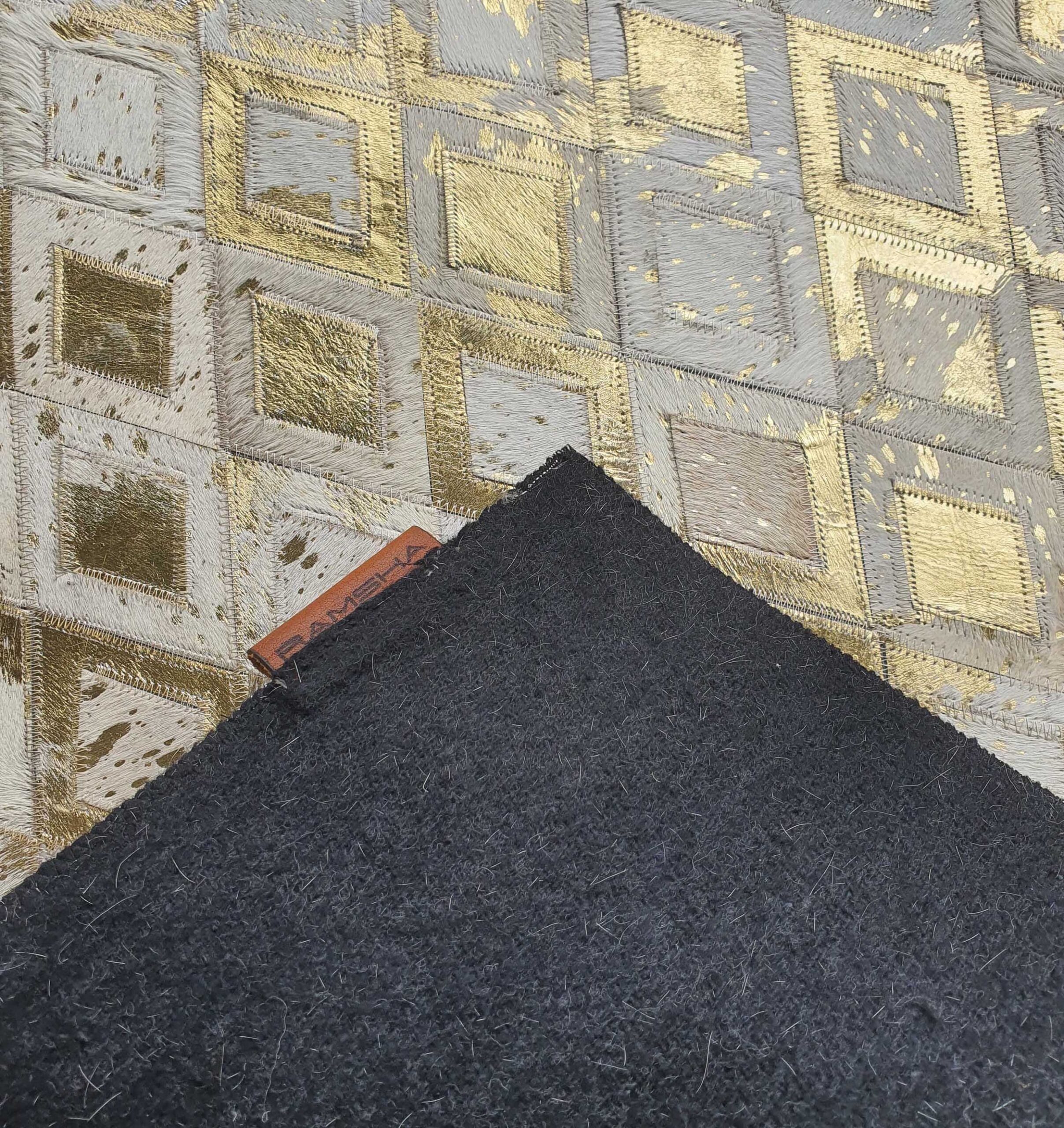 A close-up of RAMSHA HOME's Handmade Diamond Patterned Leather Rug LR-010 displays its gold and cream geometric design, partially covered in a corner by a dark gray swatch with a brown leather label.