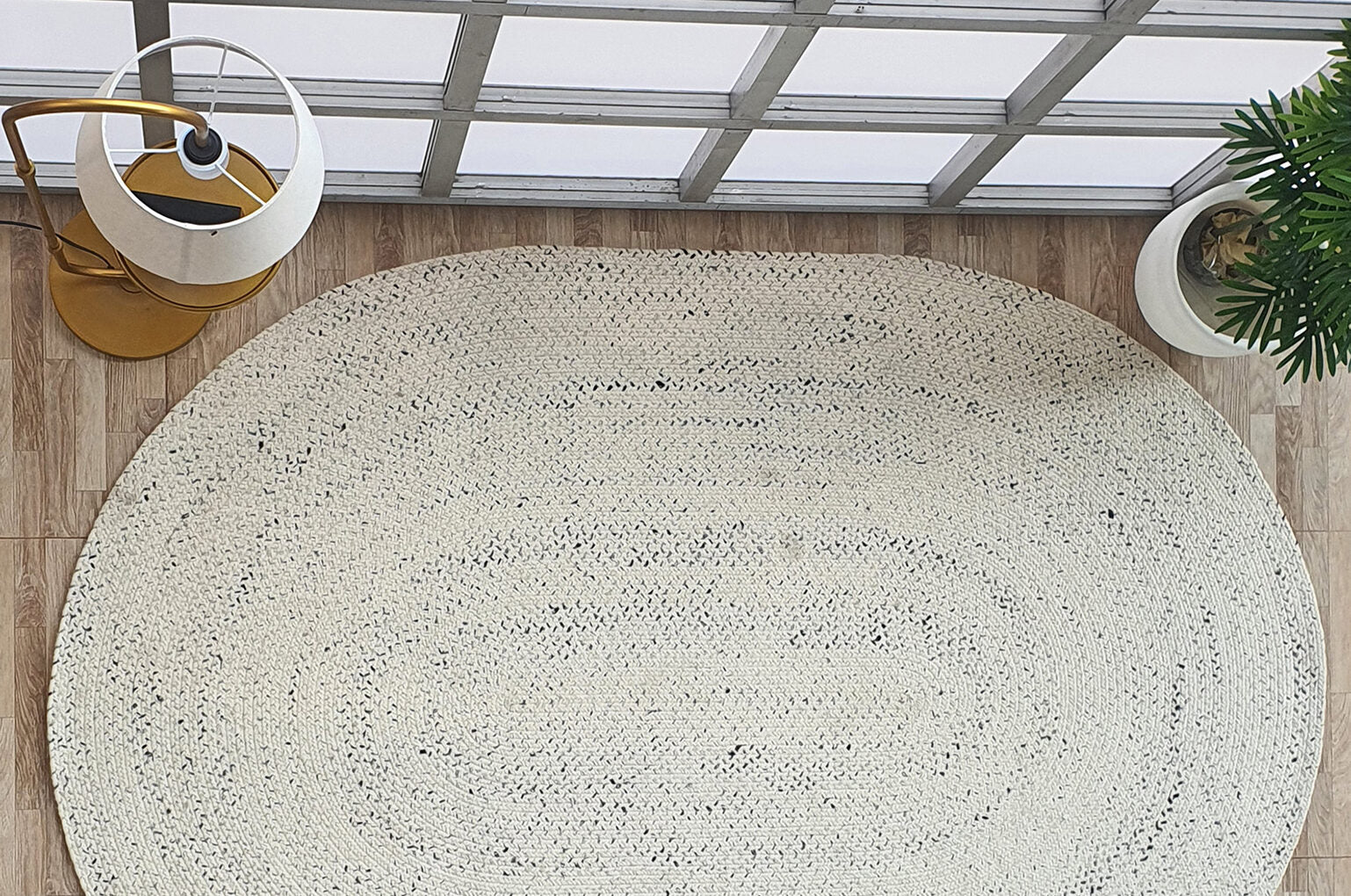 The RAMSHA HOME Amazing Handmade White & Black Color Oval Wool Floor Braided Rug - BR 047 lies on a light wood floor near a modern yellow lamp and a green plant, with large windows behind streaming in natural light.