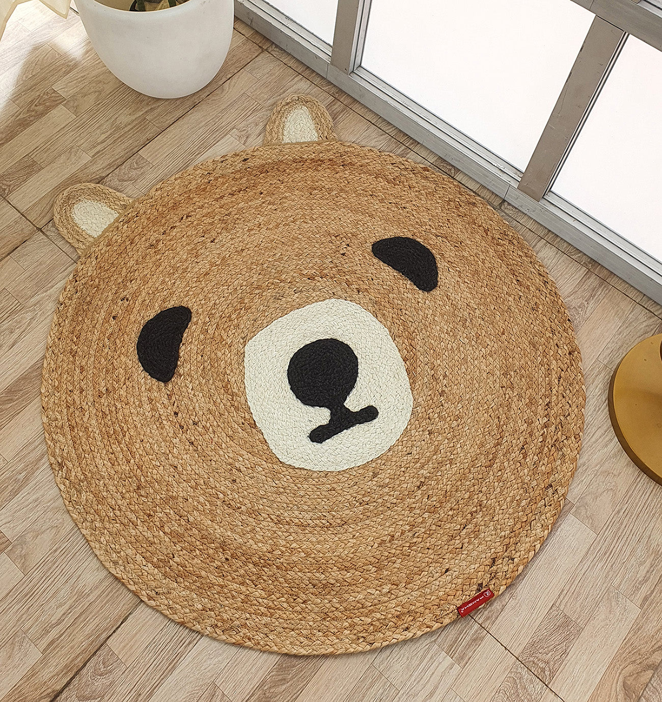 The Amazing Handmade Bear Shape Jute Rug - BR 022 (90cm) by RAMSHA HOME, with bear face details and ears in black, white, and beige, is placed on a light wood floor near a window.