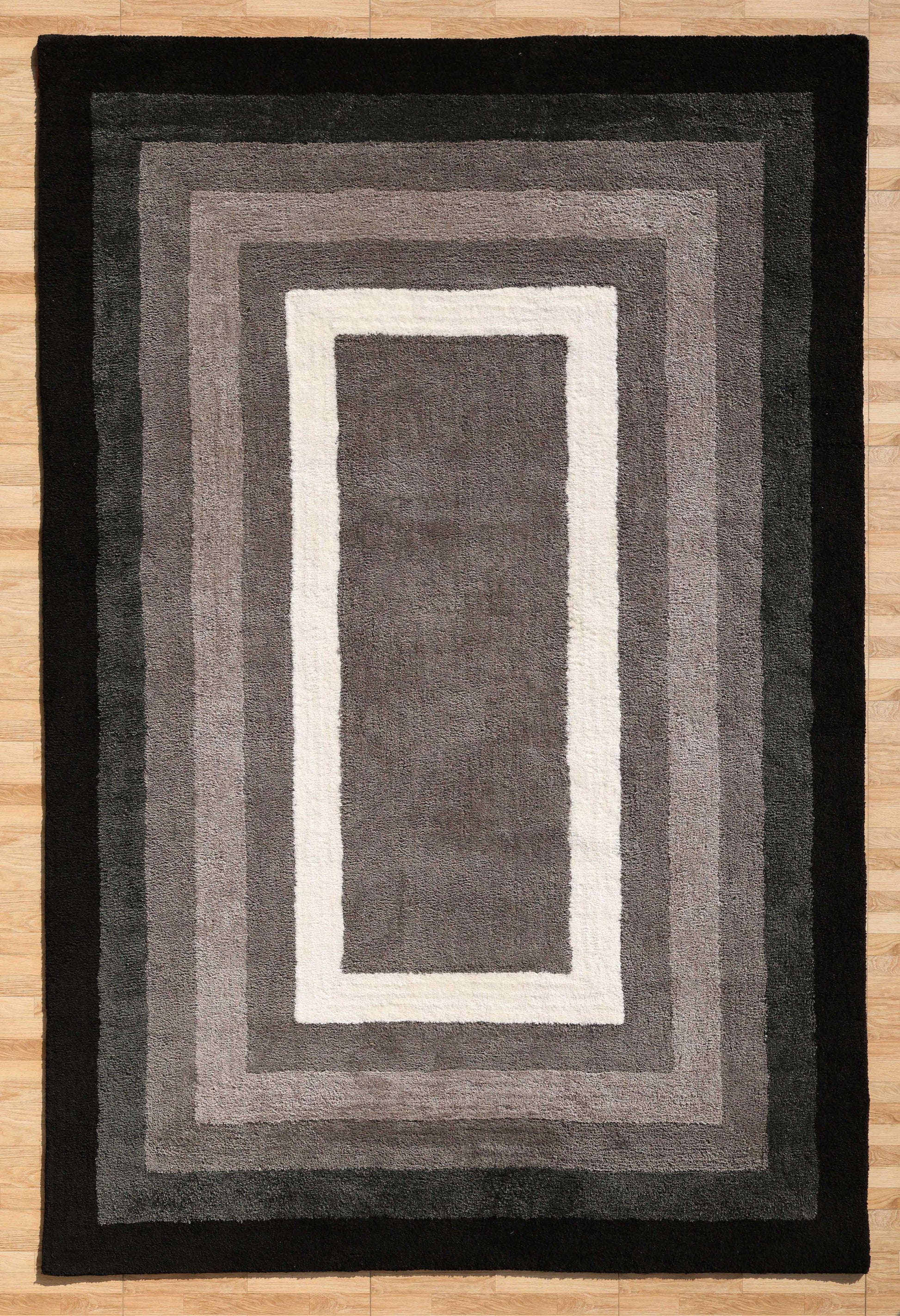 The RAMSHA HOME Black Shaggy Area Rug for Large Living Room - SG 049 features plush, concentric rectangles in gray, black, and white tones. Its soft texture and gradient design enhance comfort and depth on any light wooden floor.