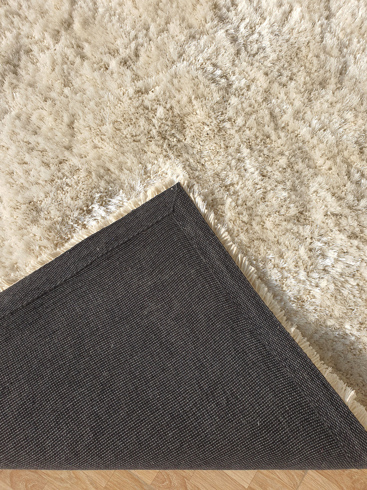 A corner of the Gold Color Shaggy Area Rug for Large Living Room - SG 03 B by RAMSHA HOME is flipped over in a dark living room, revealing the light, plush texture of the carpet beneath on a wooden floor.