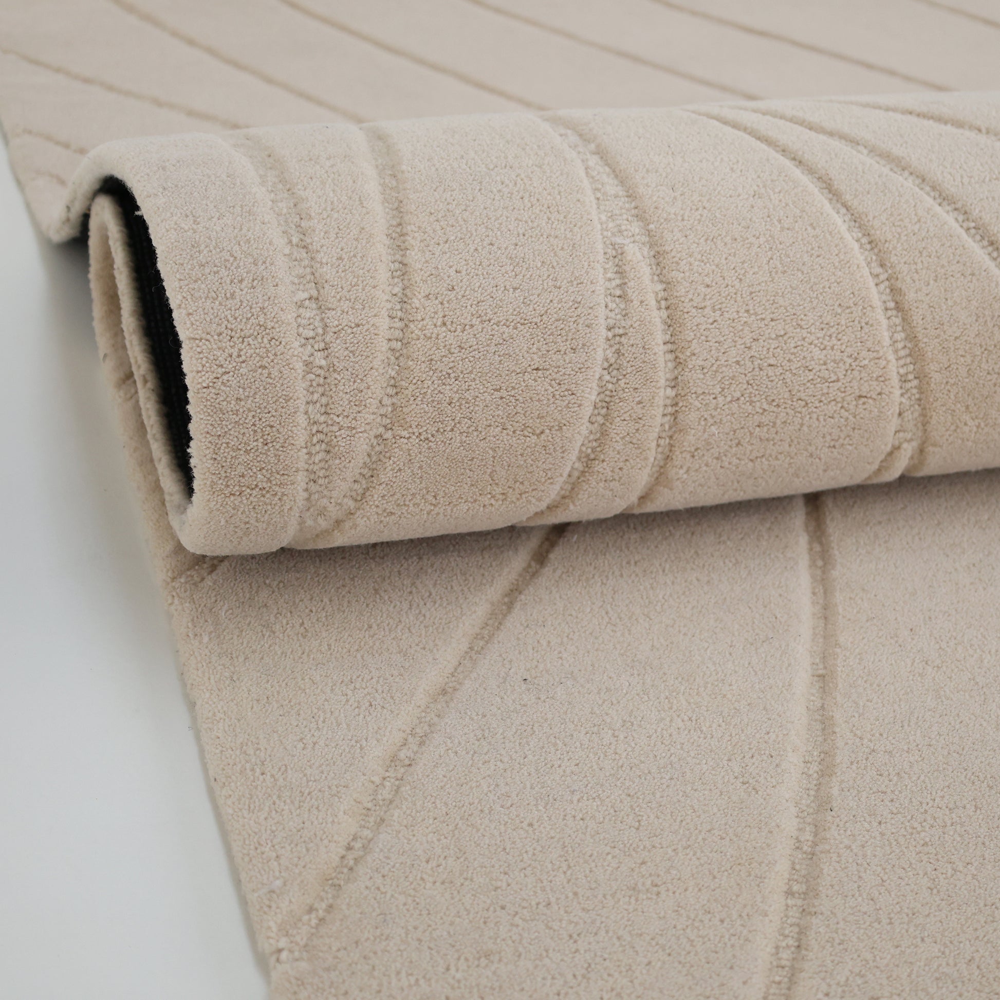 A close-up of the RAMSHA HOME Beige Color Handmade Rug - HTW 105 (300x400 cm) shows its raised, wavy carved texture and neutral wool finish, partially rolled up on a white surface.