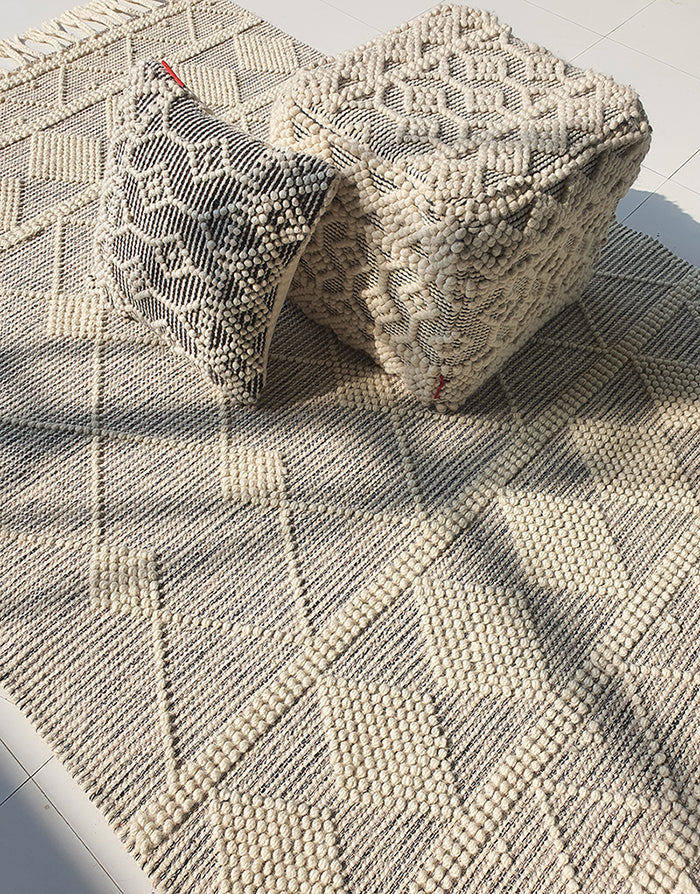 The RAMSHA HOME Fringes Woven Rug For Indoor, Floor Rug SRN-017 in grey and ivory diamond pattern is displayed with a matching cushion and cube pouf, all with raised woven designs on a sunlit white tiled floor.