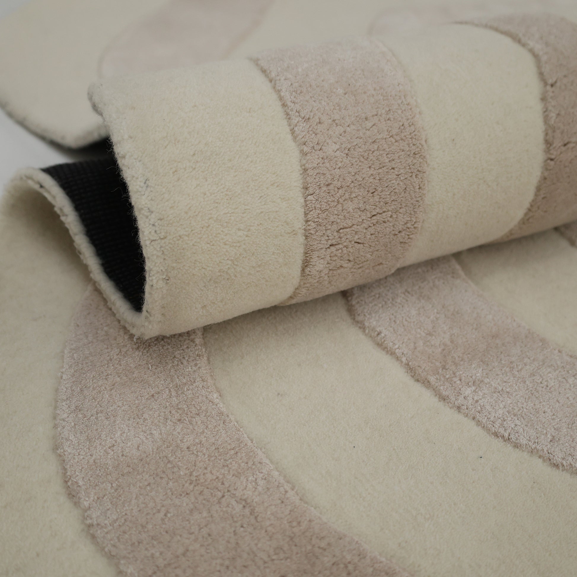 A close-up of the RAMSHA HOME Beige & Cream Wavy Handmade Area Rug – HTWV-144, hand-tufted with plush viscose and featuring beige and cream curves; partially rolled to reveal its black non-slip backing.