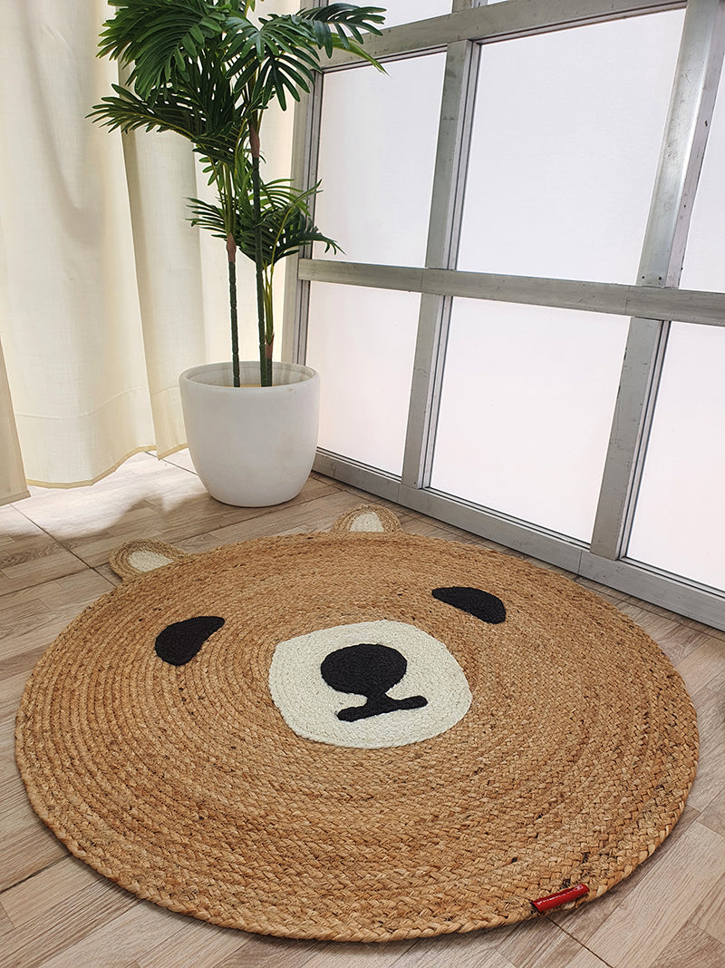 The RAMSHA HOME Amazing Handmade Bear Shape Jute Rug - BR 022 (90cm), featuring a bear face with ears, eyes, and nose, rests on a light wooden floor beside a large potted plant and a window with frosted glass panels.