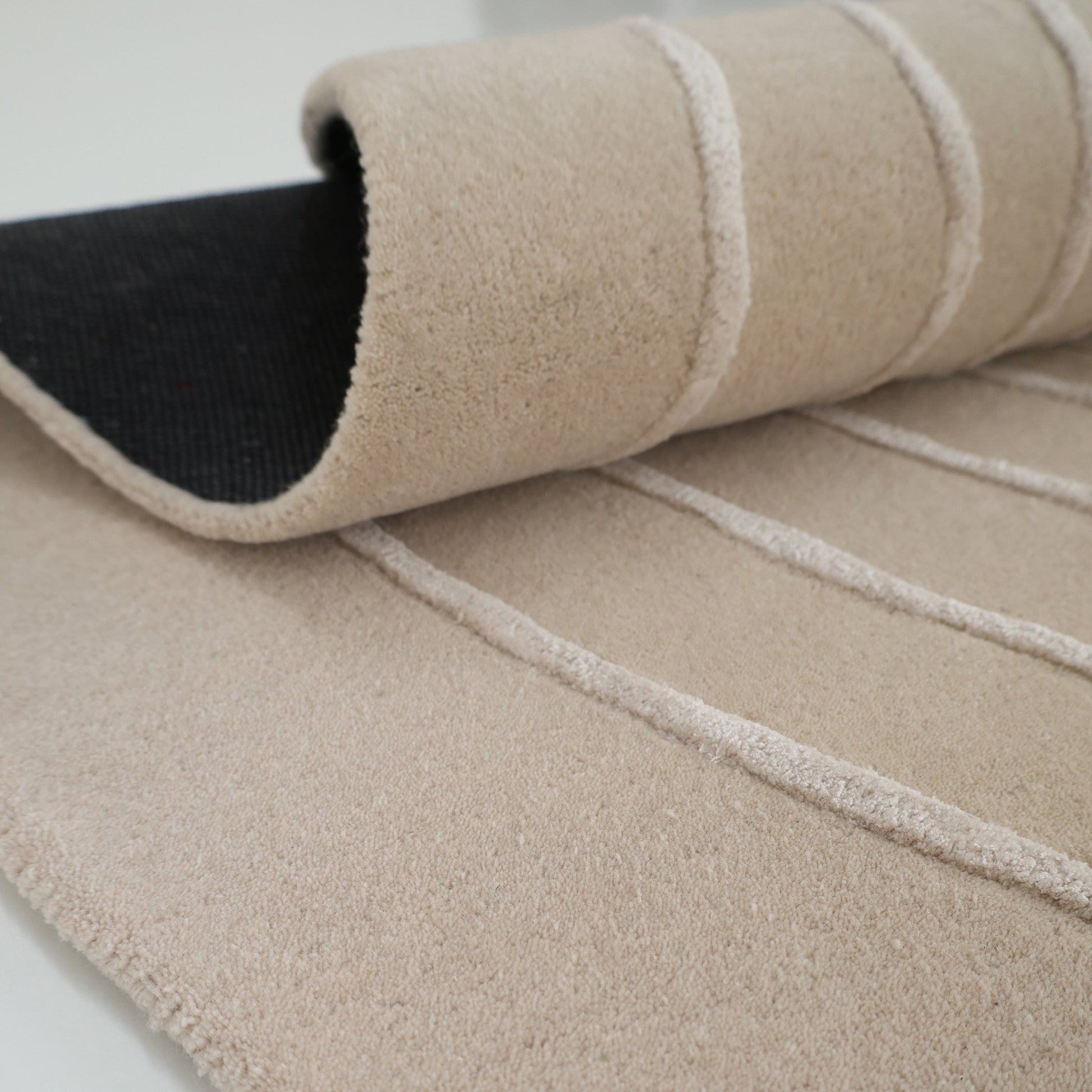 A close-up of the RAMSHA HOME Luxurious Off-White Wool & Tencel Handmade Area Rug (HTWV 110, 200x300cm), featuring a textured, hand-tufted pattern with raised lines and a partially rolled black non-slip backing.