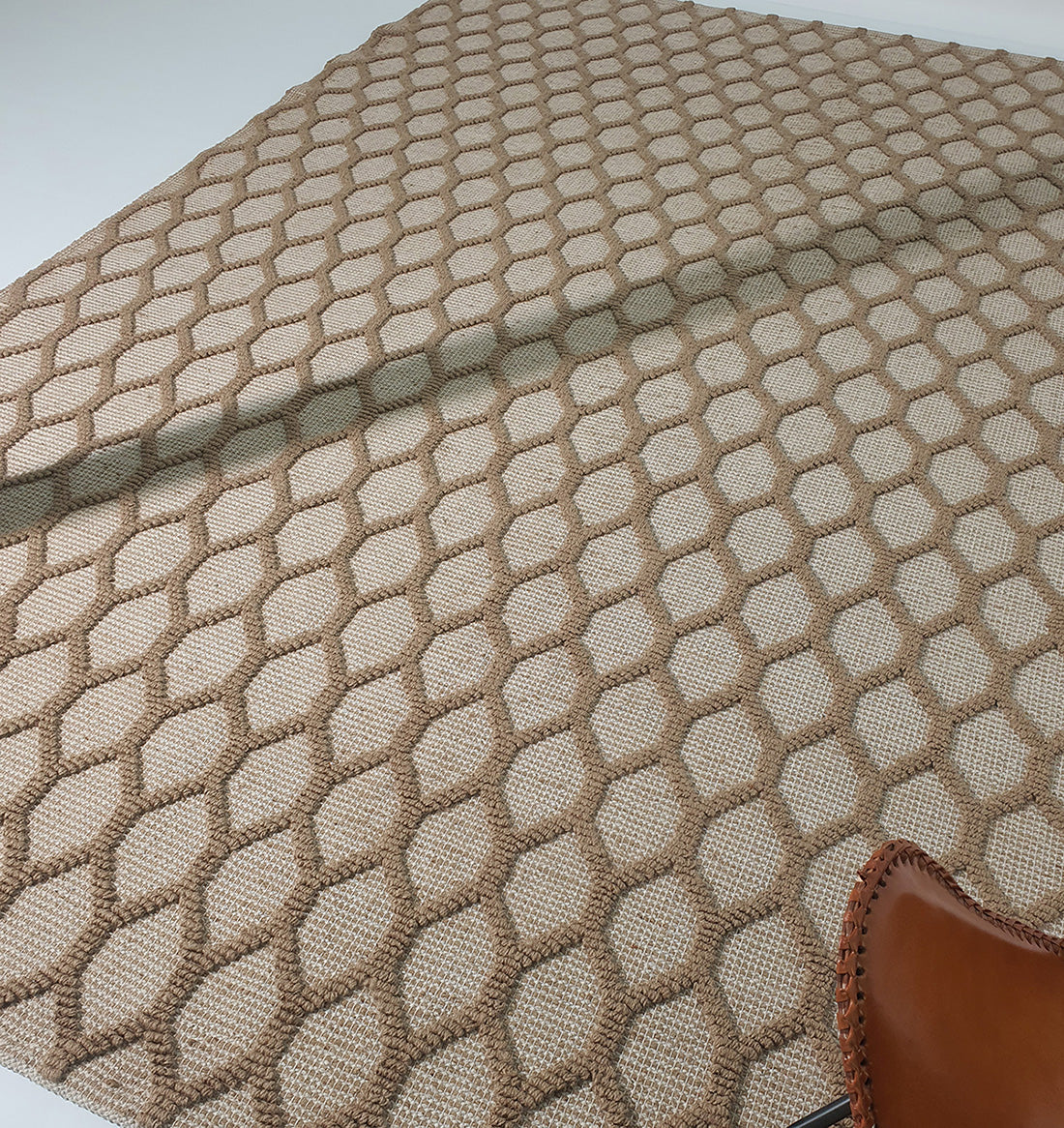 The Rectangle Woven Area Rug Neutral Beige And Brown SR-023 by RAMSHA HOME, featuring a hexagonal pattern, is partly spread out on a white surface with the corner of a brown leather chair visible in the lower right.